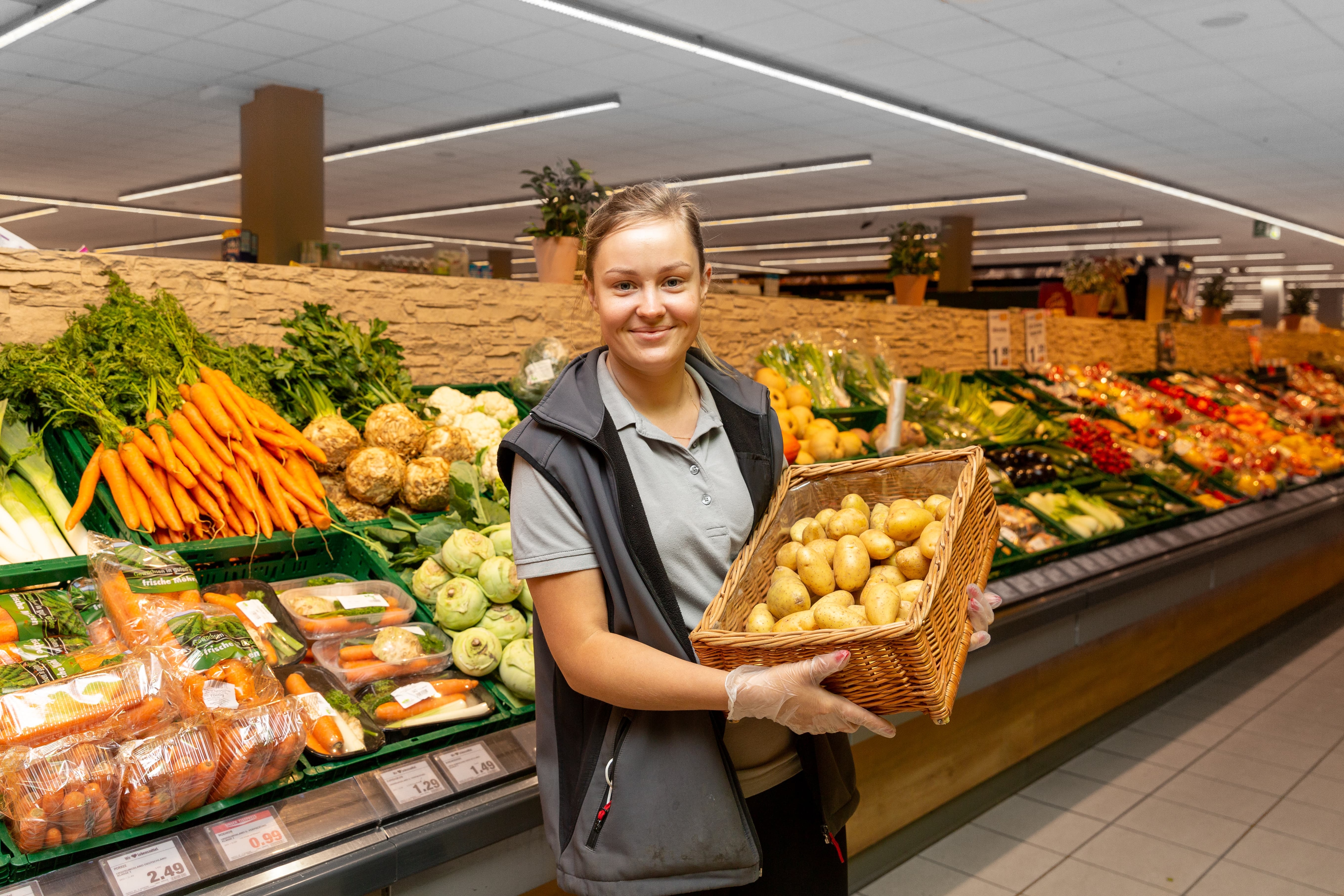 Eine lächelnde Mitarbeiterin steht in der Gemüseabteilung und hält einen Korb mit Kartoffeln in den Händen.
