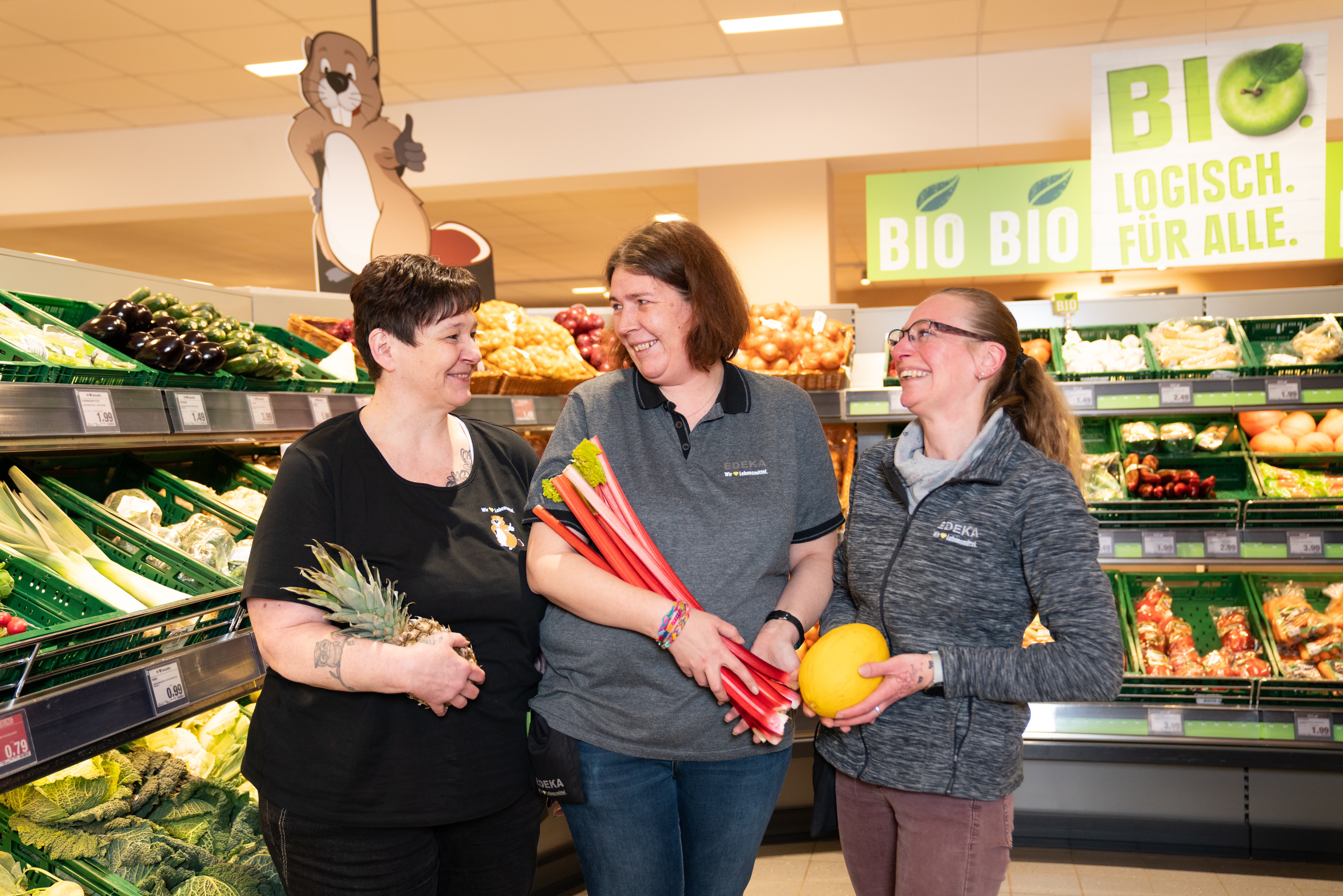 Drei weibliche Mitarbeiterinnen von EDEKA Biber stehen in der Obst- und Gemüseabteilung. In ihren Händen: Ananas, Rhabarber sowie eine Honigmelone. Alle lachen.
