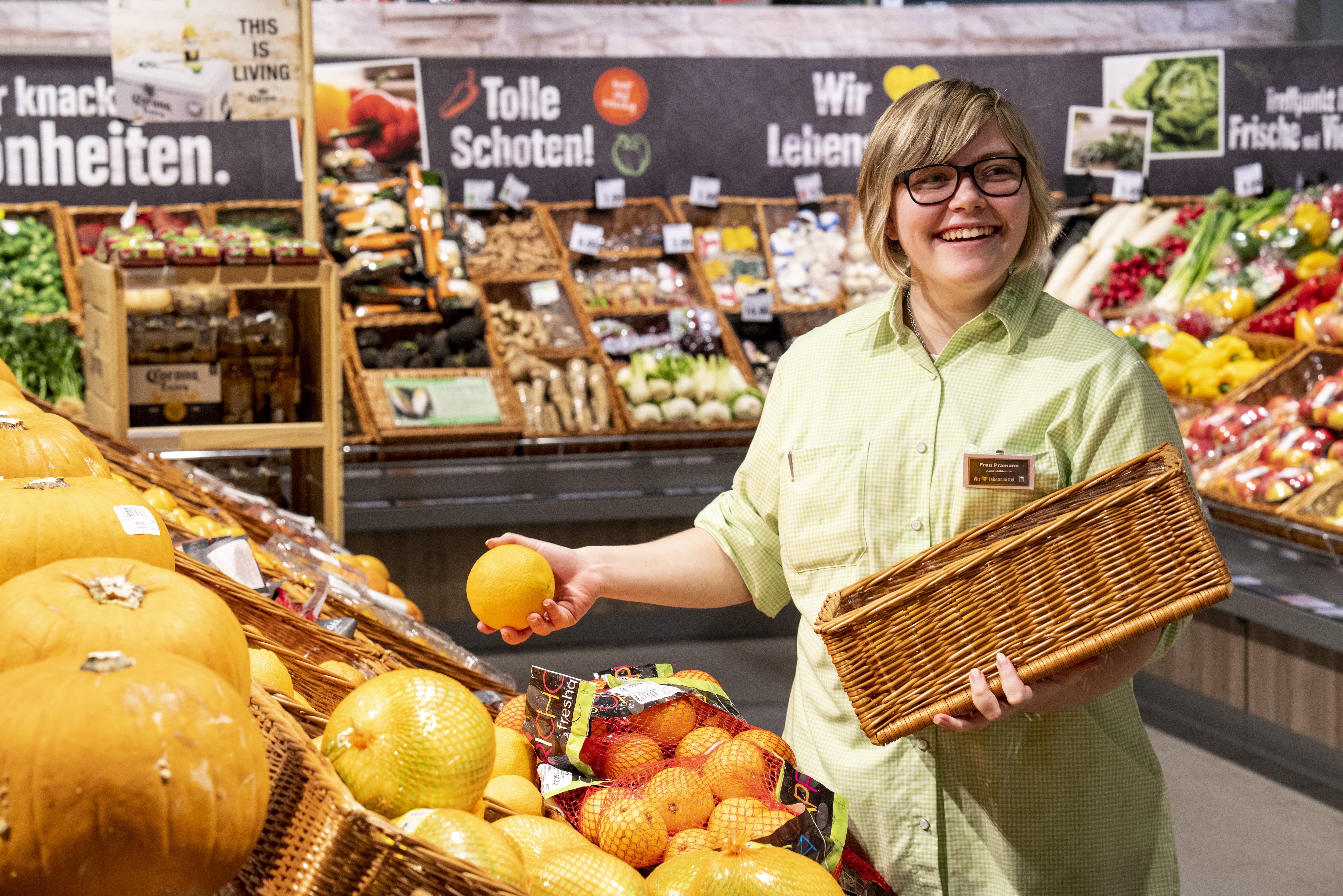 Eine lachende Auszubildende mit einem Korb im Arm legt eine Zitrusfrucht in ein Fach in der Obst- und Gemüseabteilung eines EDEKA-Markts.