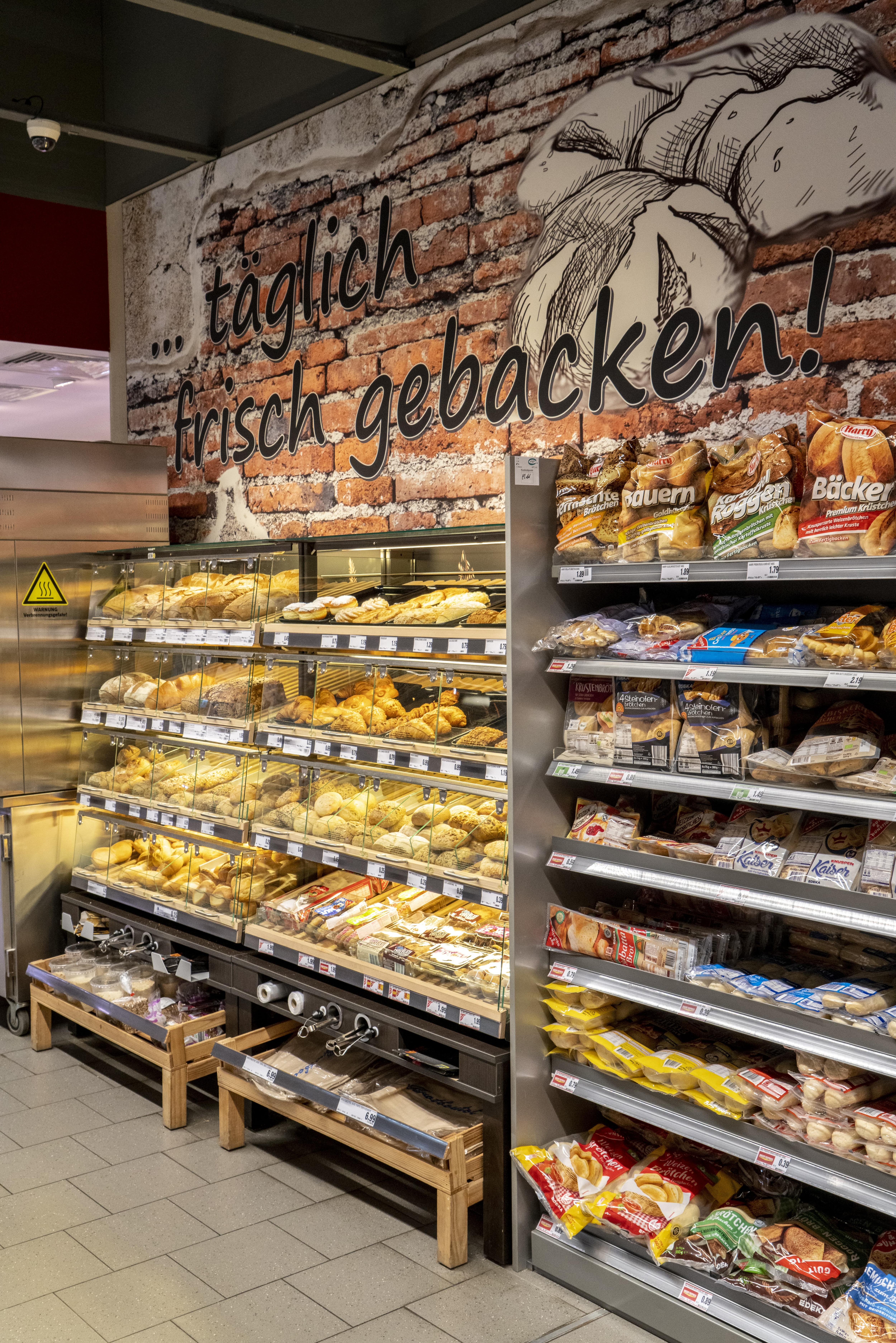 Blick auf die Brotabteilung eines EDEKA-Markts mit roten Backsteinen und der Aufschrift „Täglich frisch gebacken!“ an der Wand im Hintergrund.
