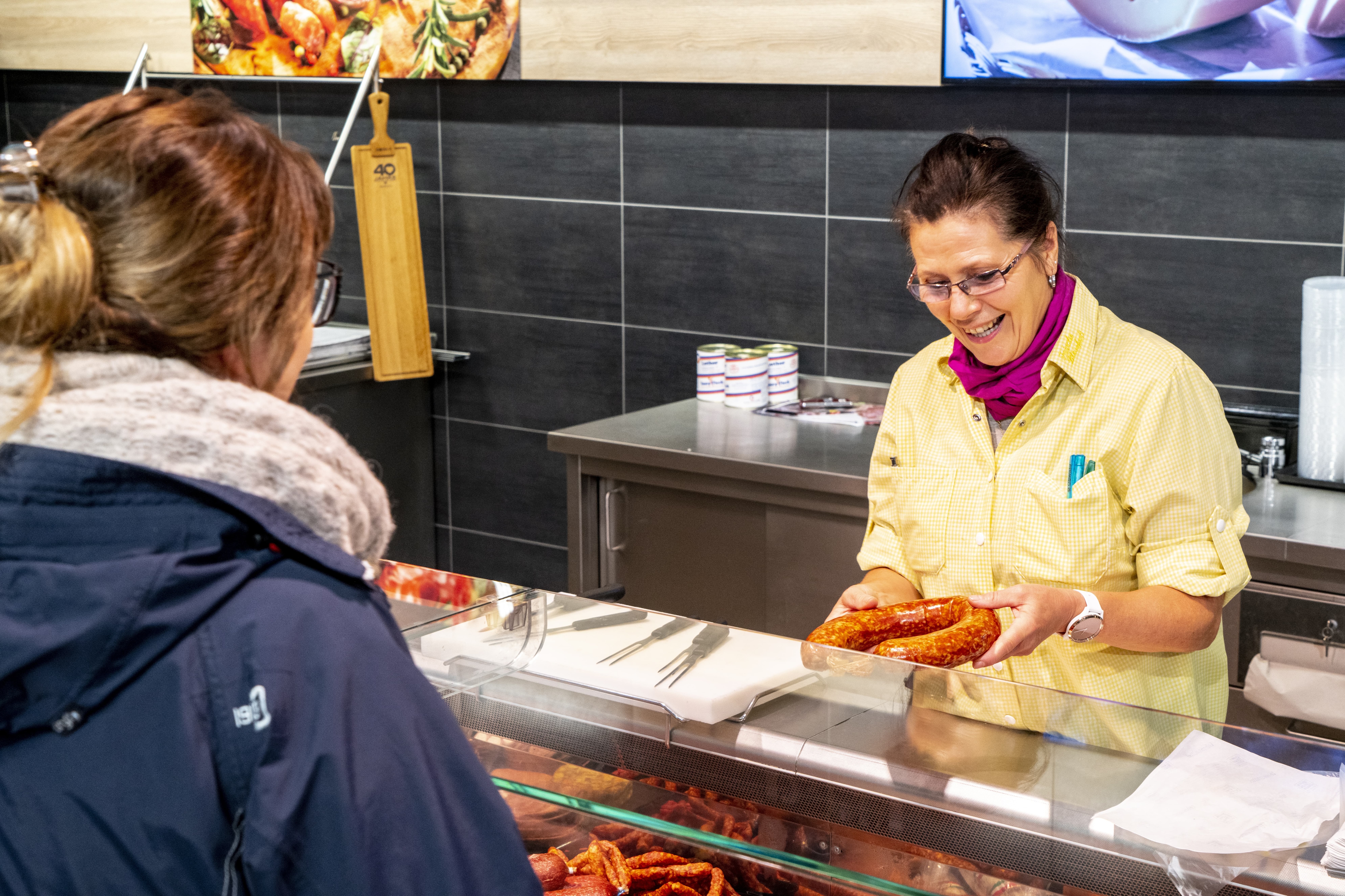 Eine EDEKA-Mitarbeiterin hält lachend eine Wurst in der Hand, während ihr an der Fleischtheke eine Kundin gegenübersteht.