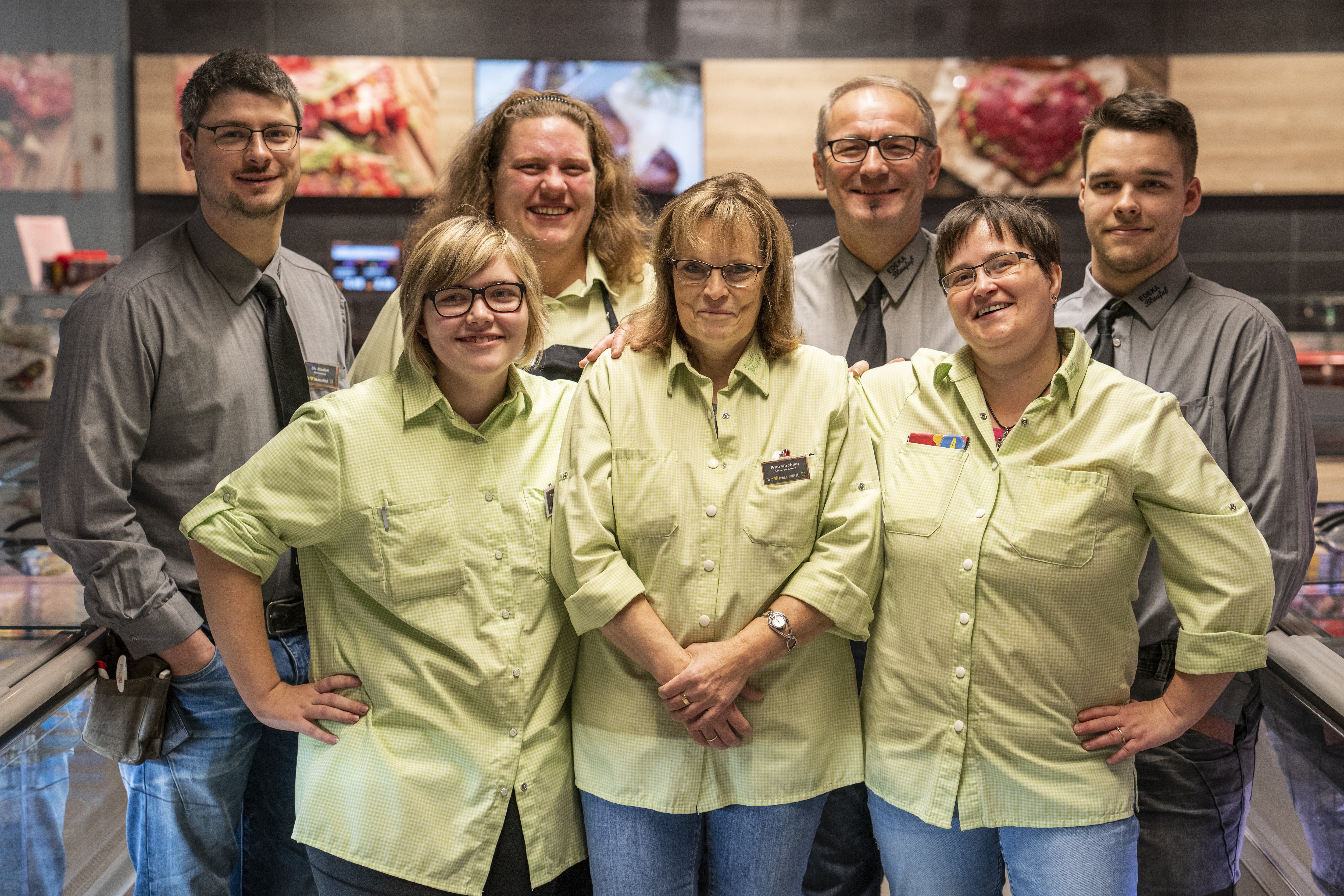 Gruppenfoto des Teams dieses EDEKA-Markts, aufgenommen vor der Frischetheke. Die Mitarbeitenden lächeln und tragen Grau oder Hellgrün.