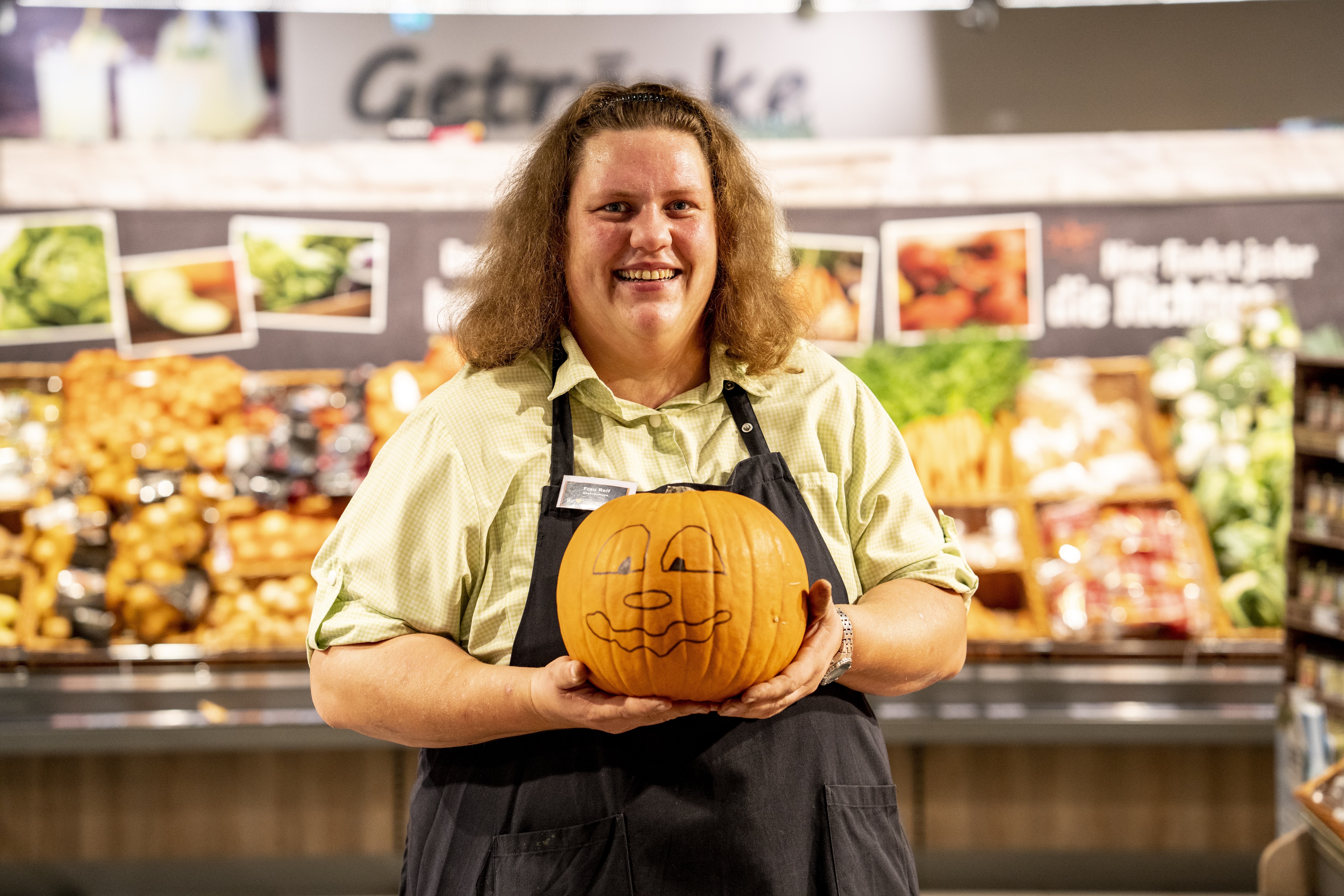 Eine Mitarbeiterin mit Schürze hält einen Kürbis hoch, auf diesem ist ein Gesicht aufgemalt.