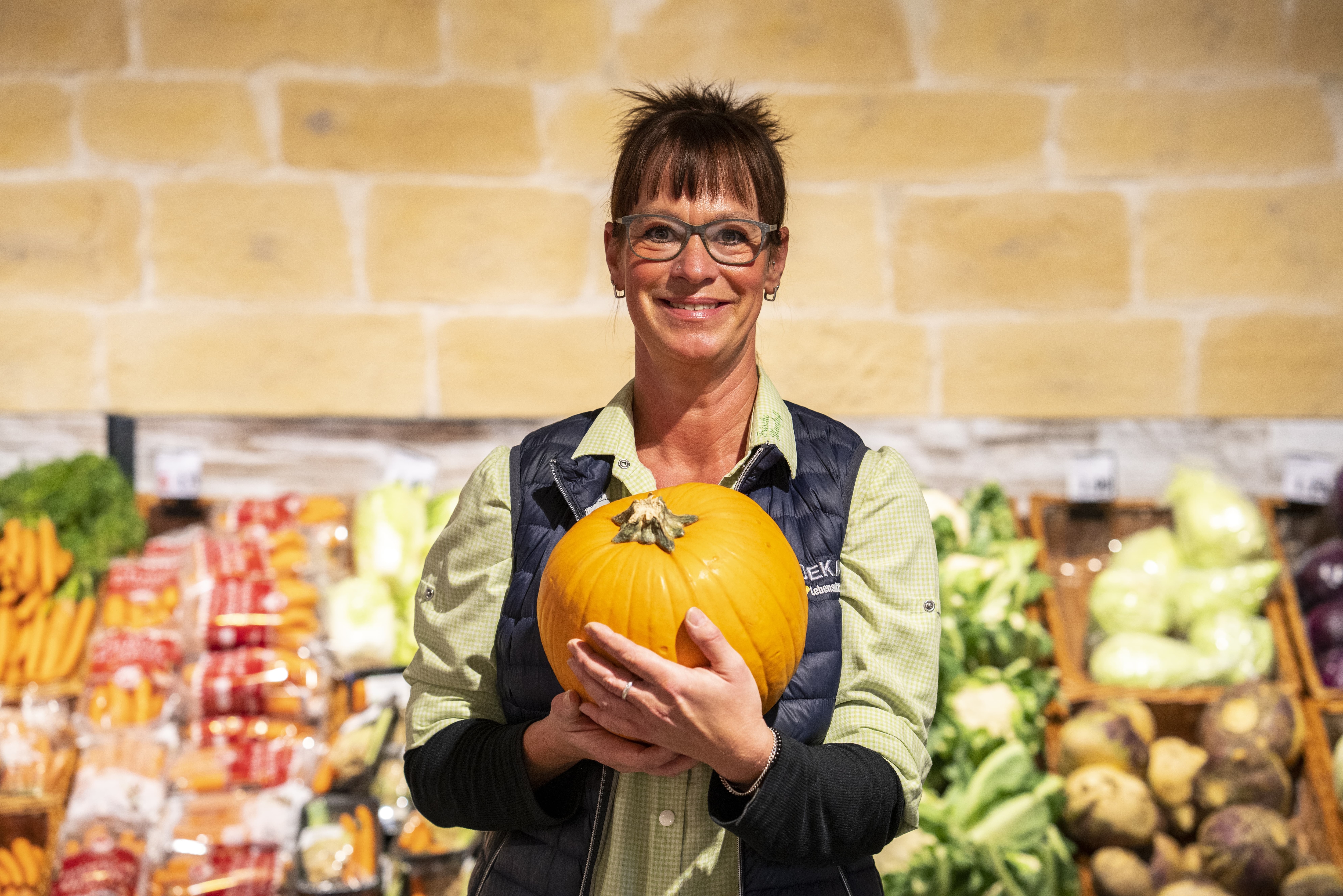 Eine lächelnde Mitarbeiterin steht in der Gemüseabteilung eines EDEKA-Marktes und hält einen Kürbis in den Armen.