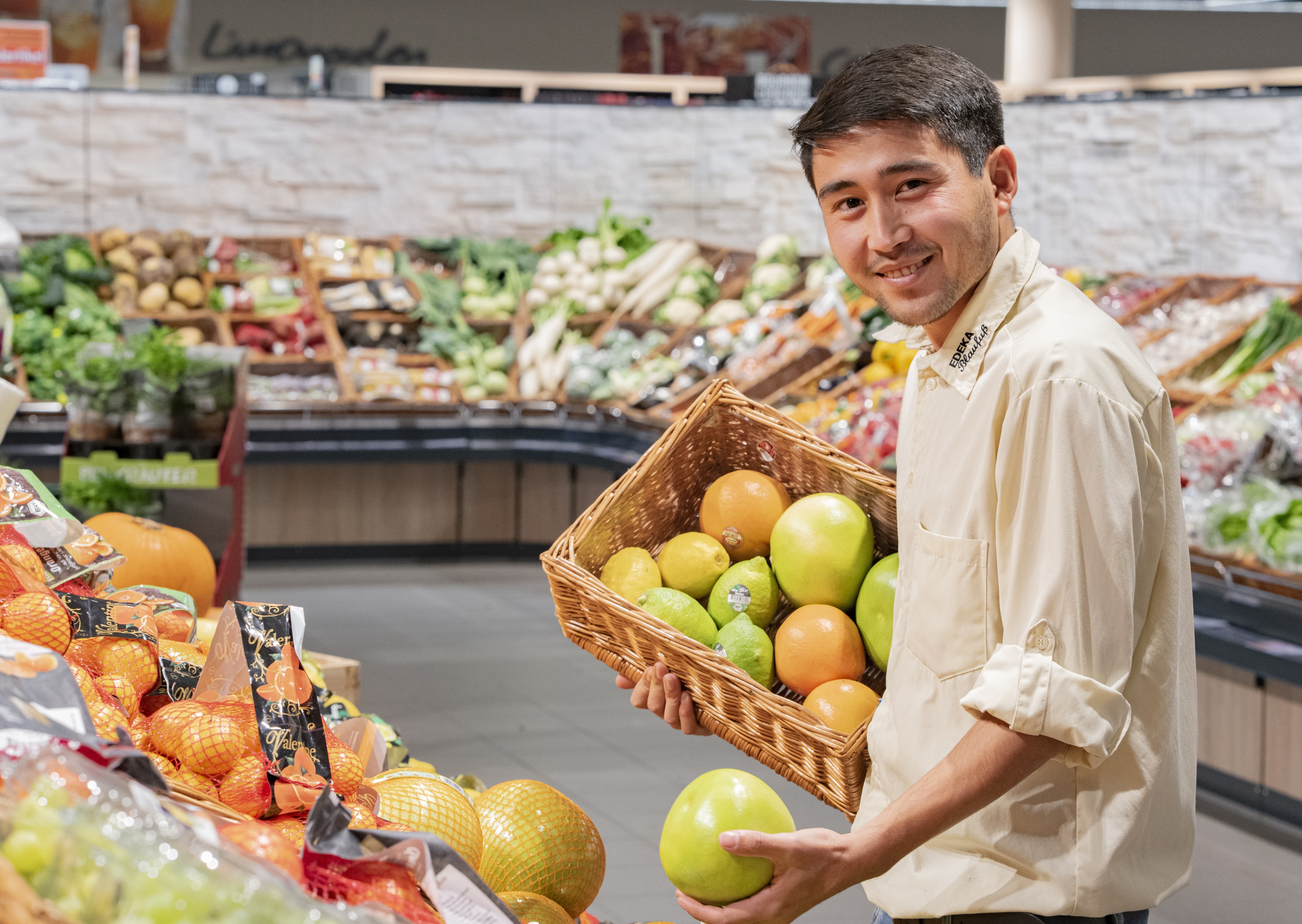 Ein Auszubildender hält einen Korb mit verschiedenen exotischen Früchten und lächelt in die Kamera. Er steht in der Obst- und Gemüseabteilung eines Edeka-Marktes.