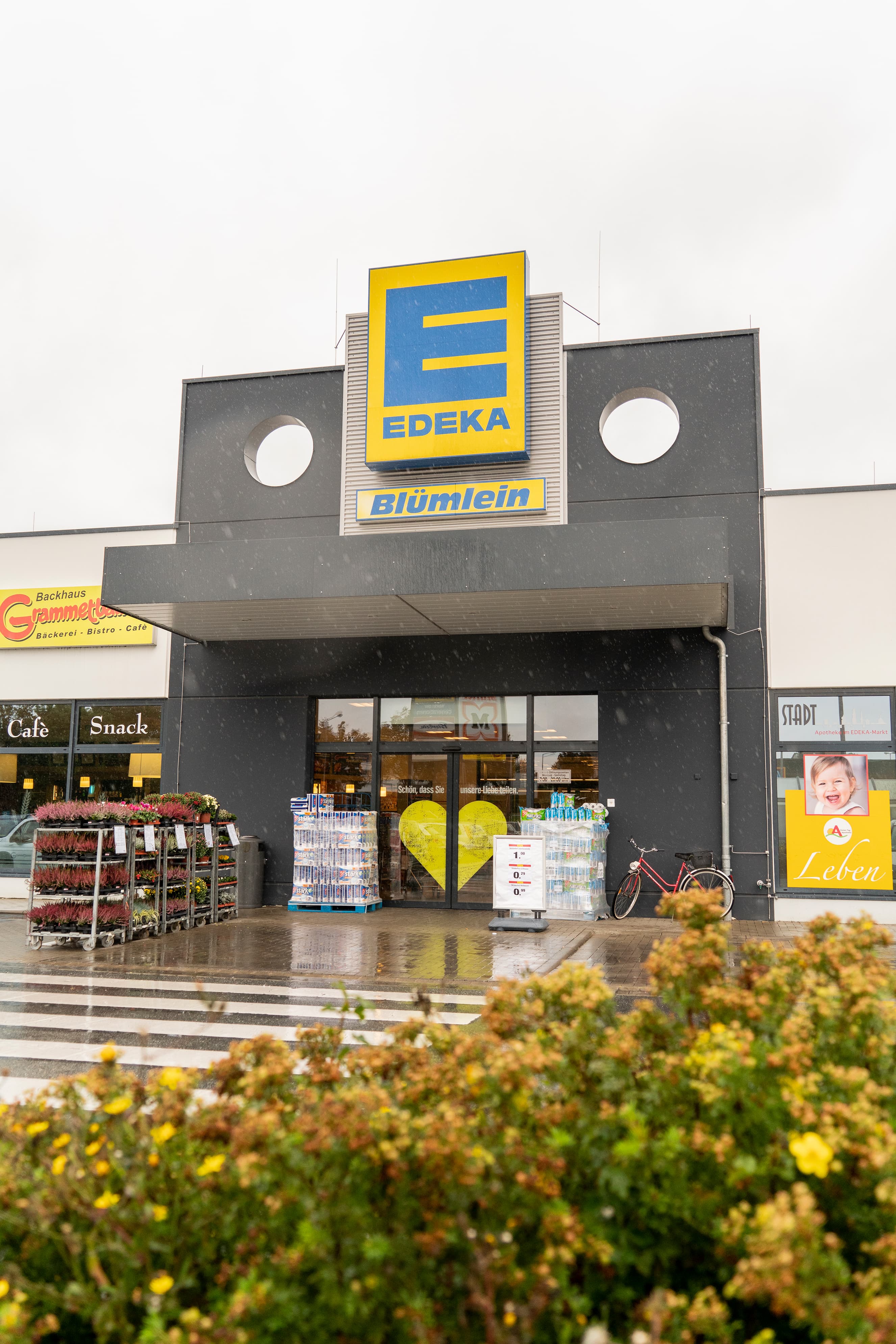Front des Markts, bei regnerischem Wetter, mit dem Schriftzug EDEKA Blümlein über dem Eingang und Büschen im Vordergrund.
