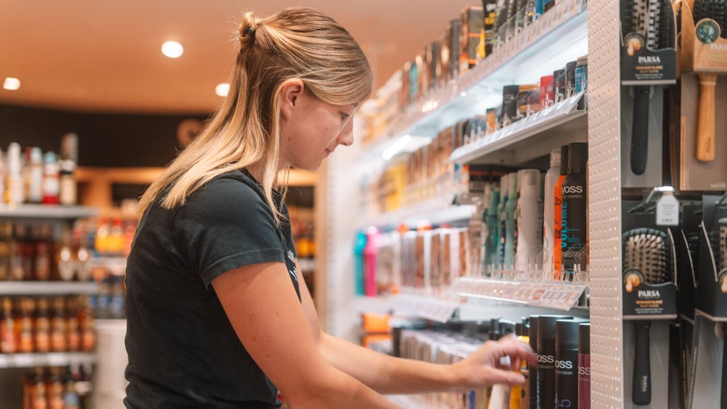 Eine Mitarbeiterin im Profil am Drogerieregal, die nach den Haarsprays greift.