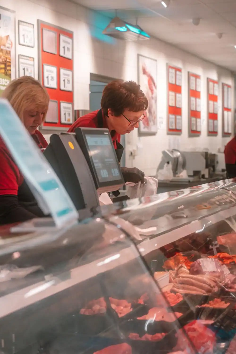 Zwei Mitarbeiterinnen hinter der Frischetheke beim Fleisch und bedienen Kunden.