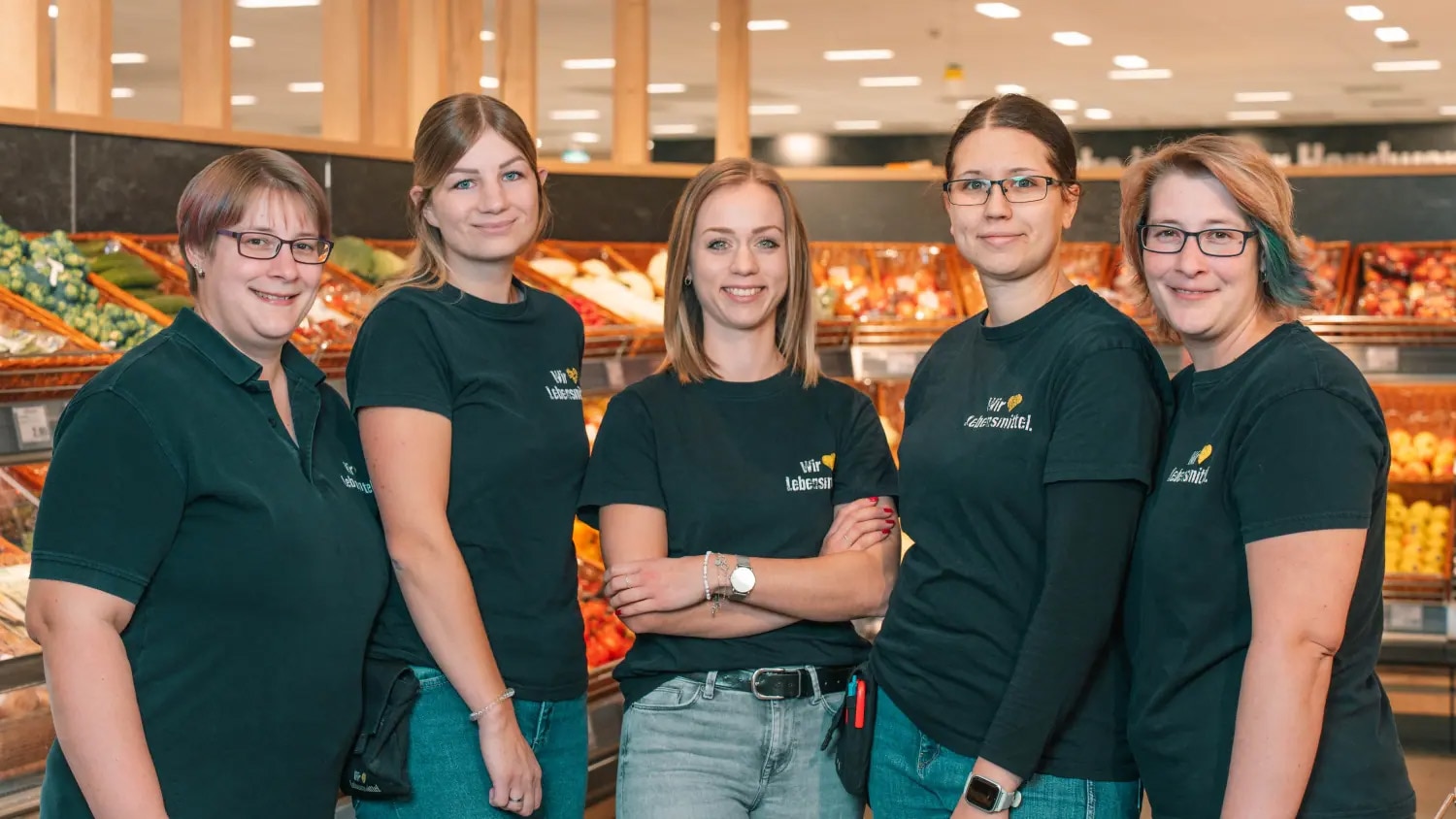Fünf freuen lächeln in die Kamera und stehen in der Obst- und Gemüseabteilung. Sie tragen Mitarbeiter-Tshirts.