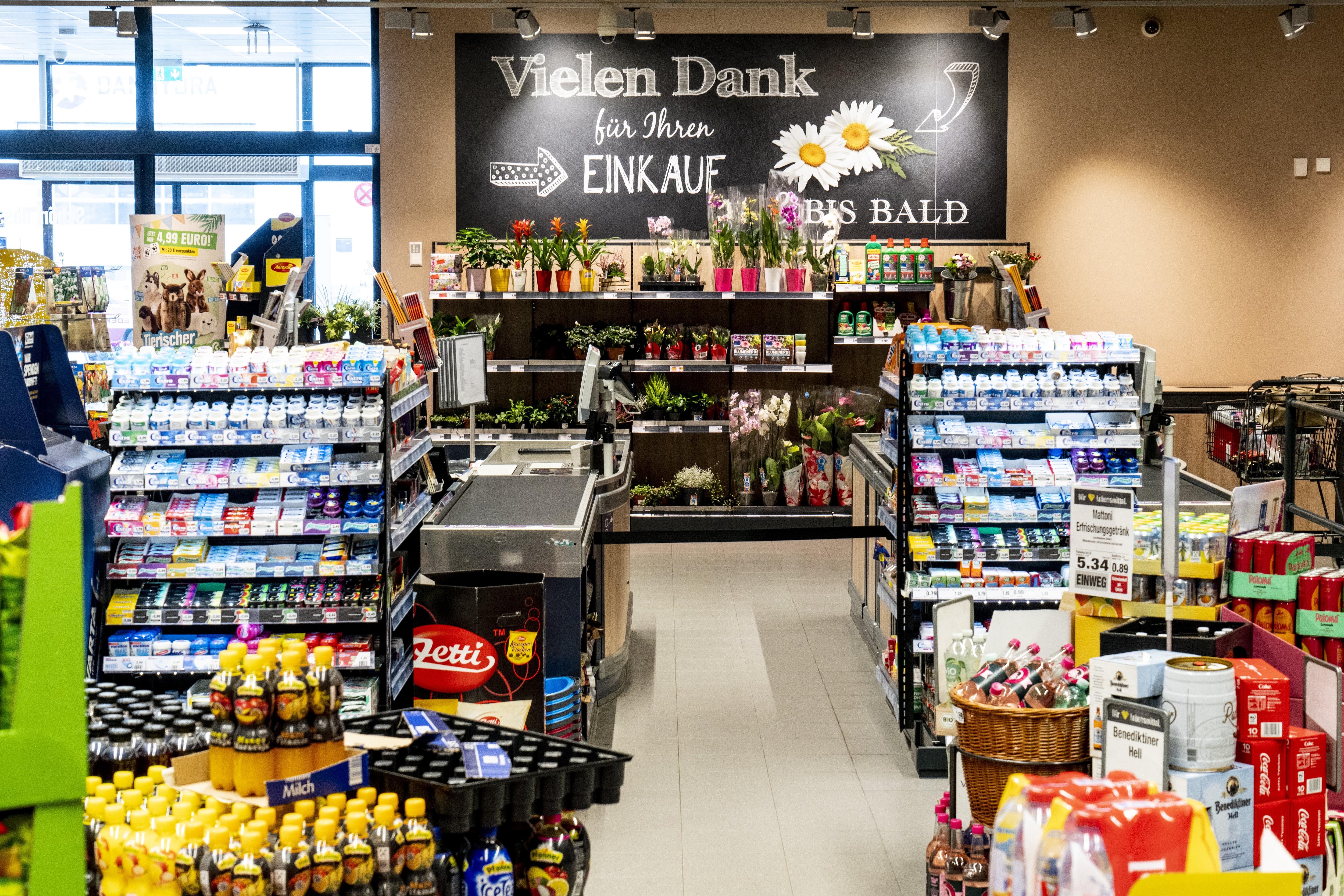 Der Kassenbereich eines EDEKA mit reich und bunt bestückten Produktregalen im Vordergrund und dem Schild „Vielen Dank für Ihren Einkauf. Bis bald.“ im Hintergrund.