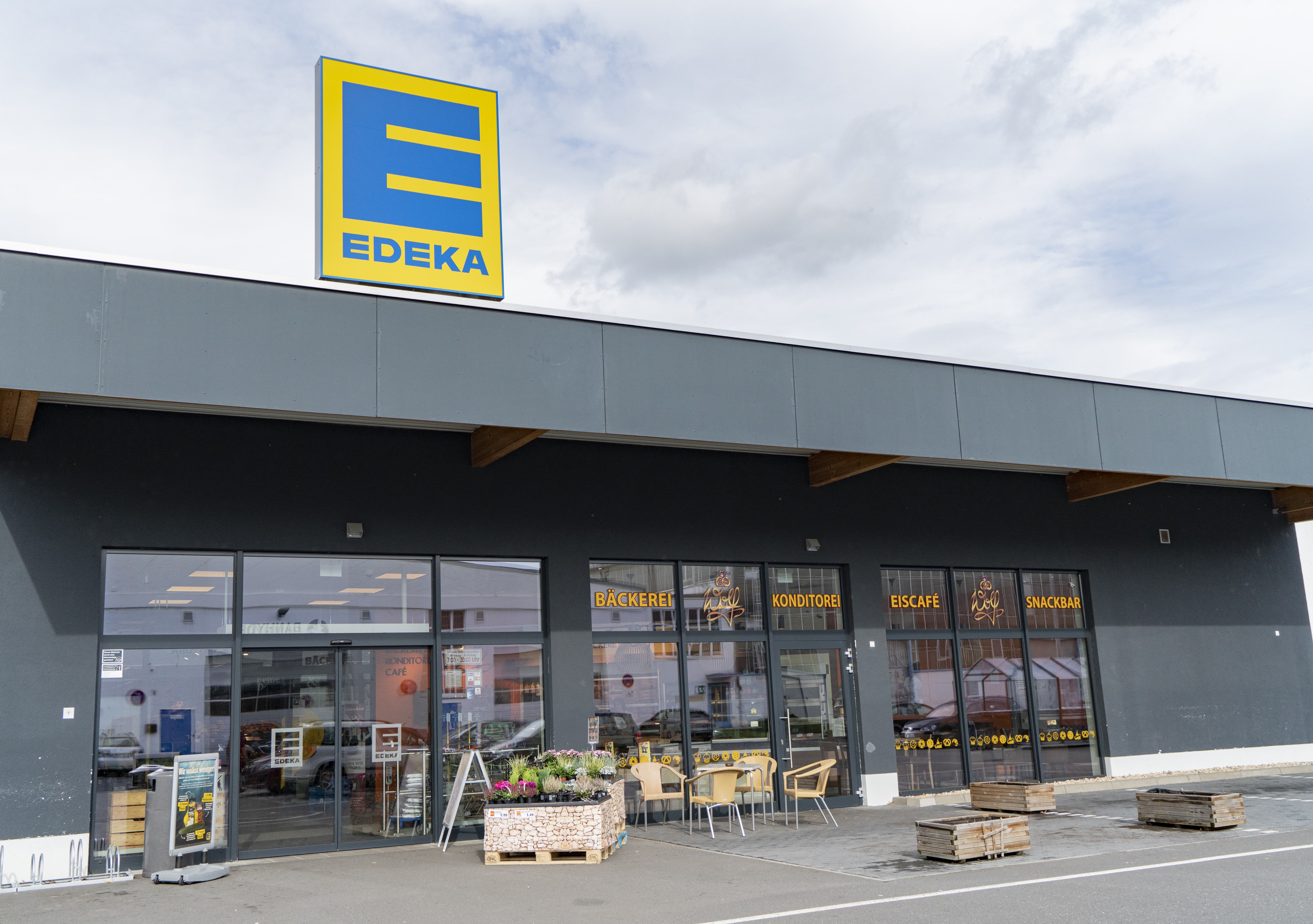 Front des Markts, sonnenbeschienen, mit dem Schriftzug EDEKA über dem Eingang. Es gibt eine Bäckerei, eine Konditorei, ein Eiscafé und eine Snackbar, den Schriftzügen auf den Fenstern zufolge.