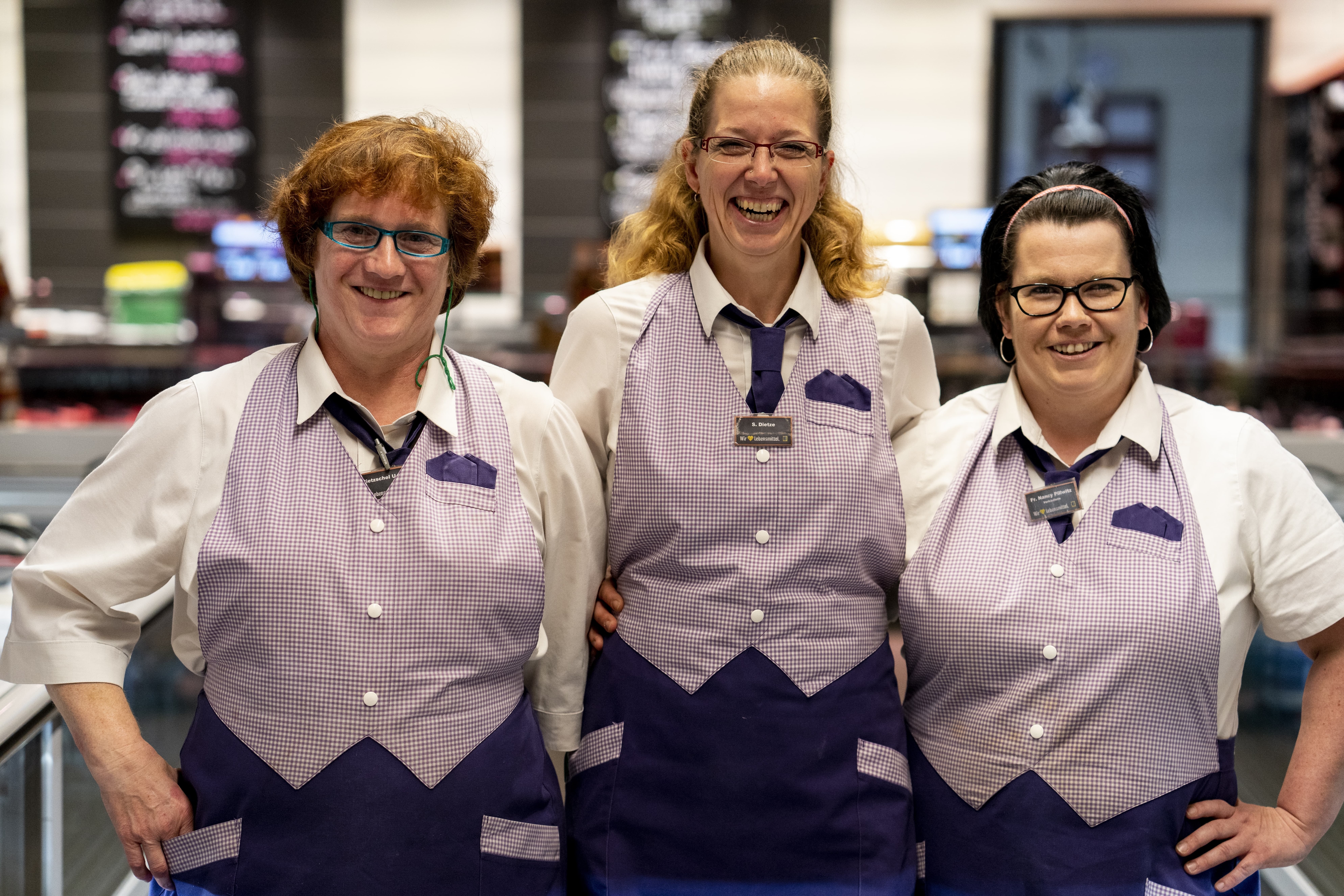 Drei Mitarbeiterinnen lachen in die Kamera. Sie tragen eine Uniform aus weißem Hemd, lila Krawatte und einer Schürze, die oben aussieht wie eine lila-weiß-karierte Weste.