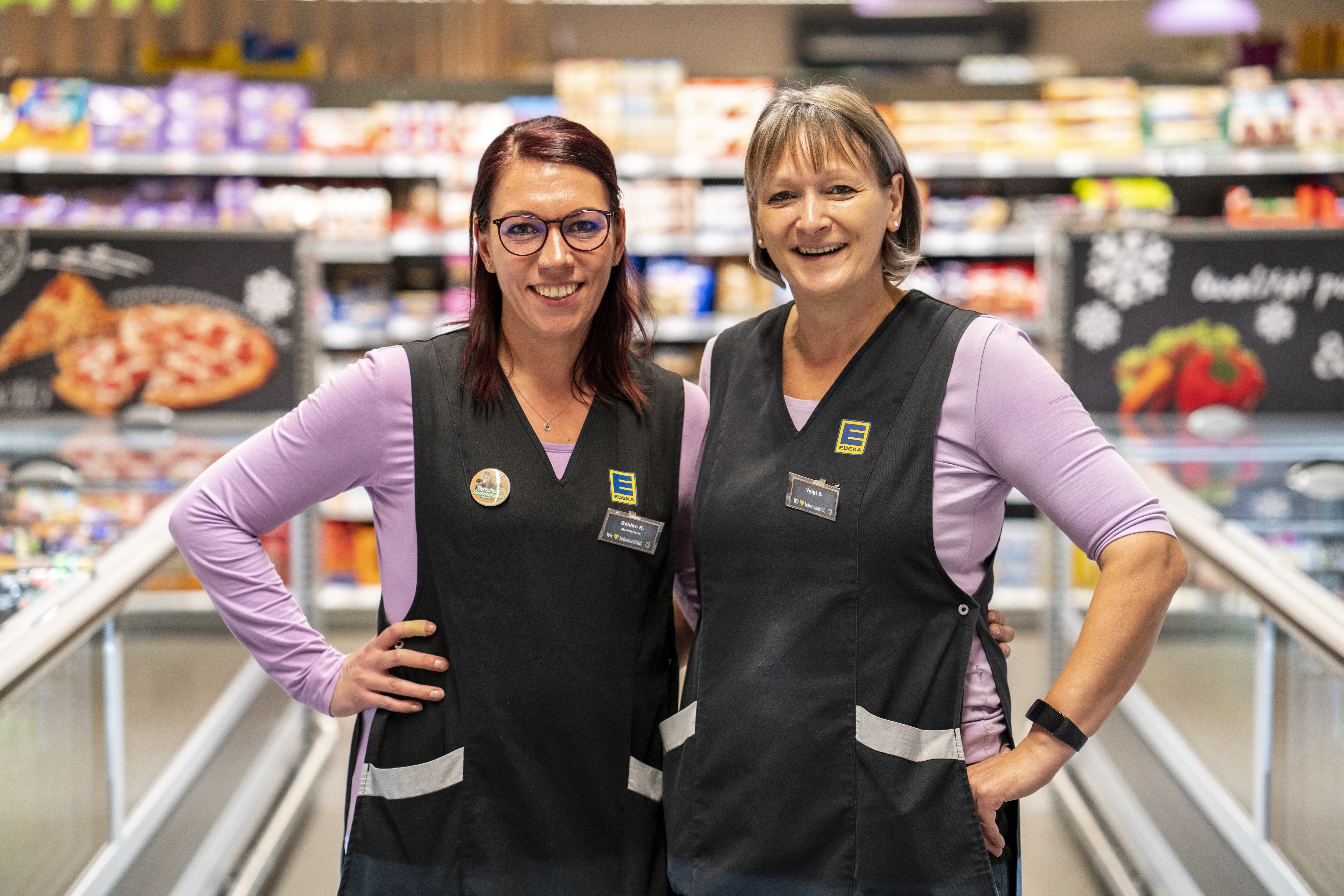 Zwei Mitarbeiterinnen stehen Arm in Arm und lächeln in die Kamera, im Hintergrund Produktregale.