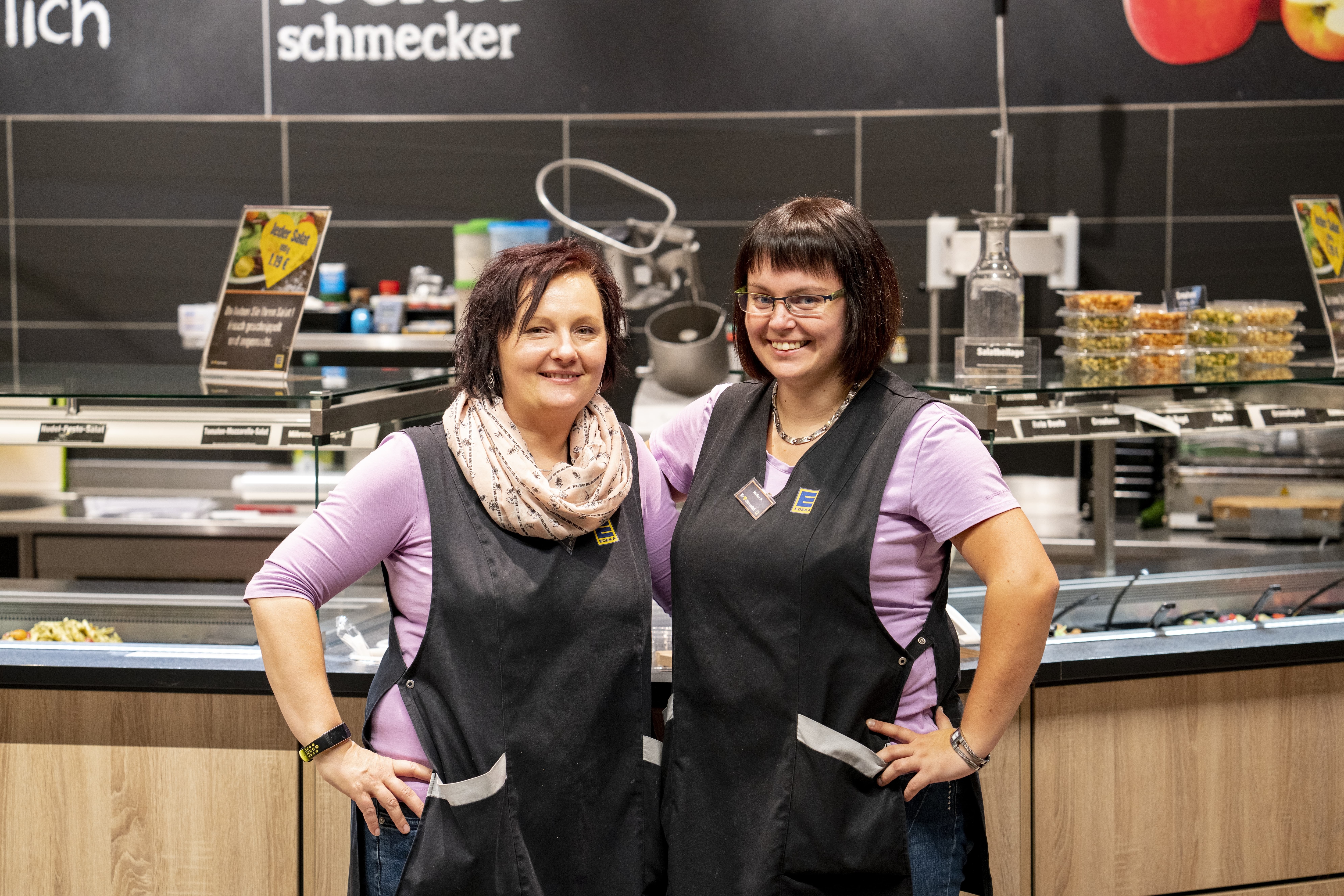 Zwei Mitarbeiterinnen stehen Arm in Arm und lächeln in die Kamera, im Hintergrund eine Bedientheke..