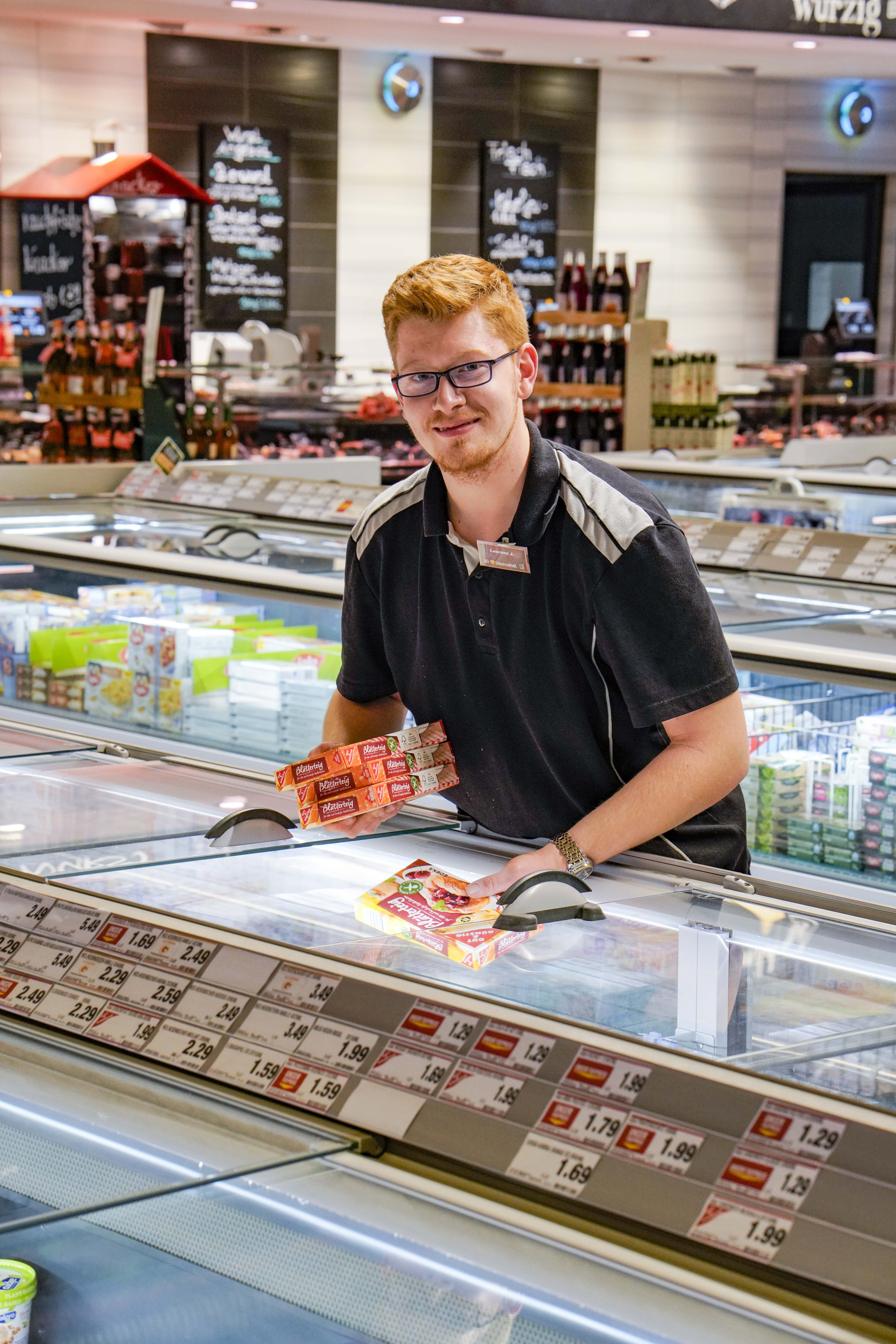 Ein lächelnder Mitarbeiter schaut in die Kamera, während er Produkte in eine Tiefkühltruhe in einem EDEKA-Markt einordnet.