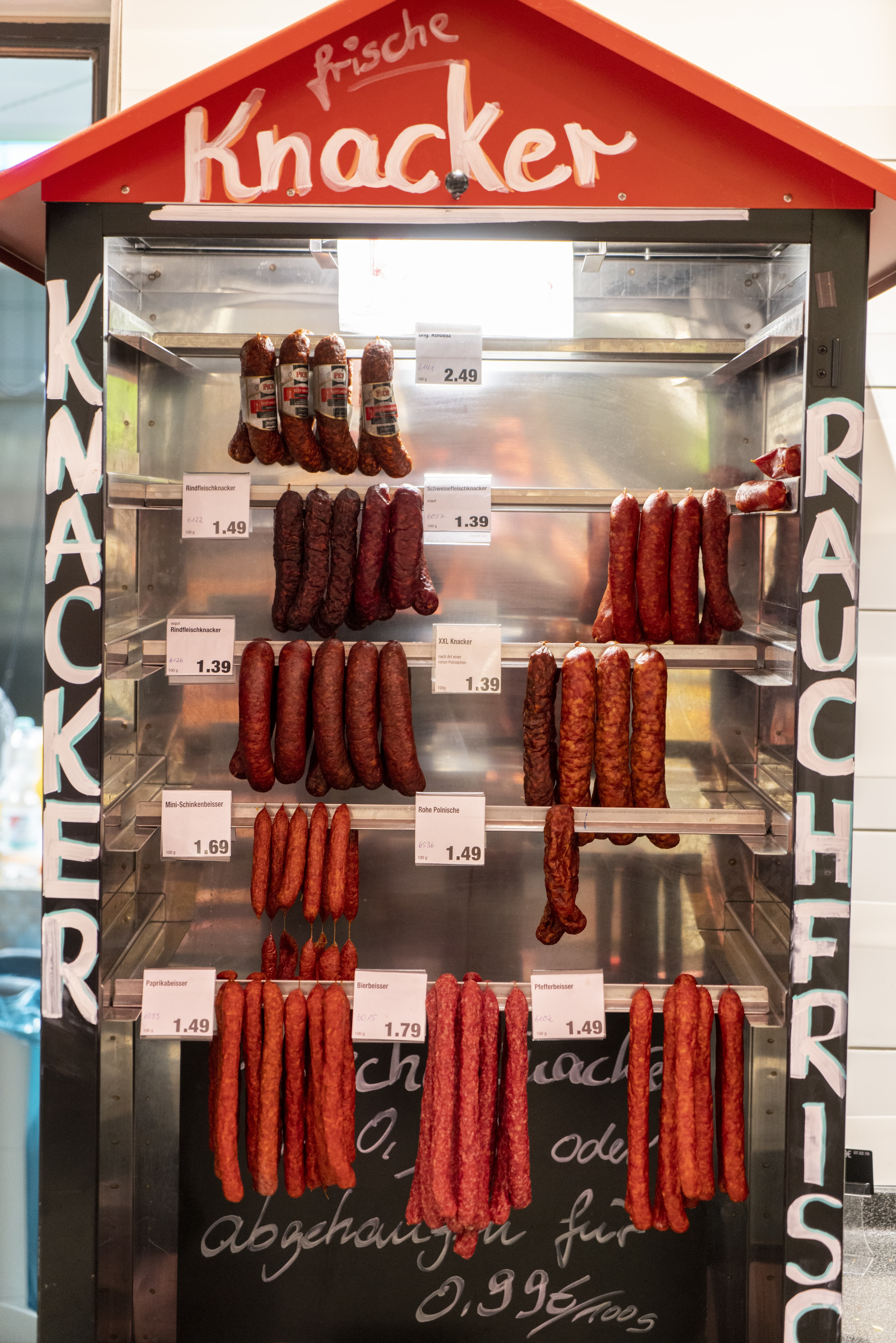 Eine Art Regal mit aufgesetztem rotem „Hausdach“, in dem Würste hängen. Auf dem Regal steht: „Frische Knacker. Knacker rauchfrisch.“
