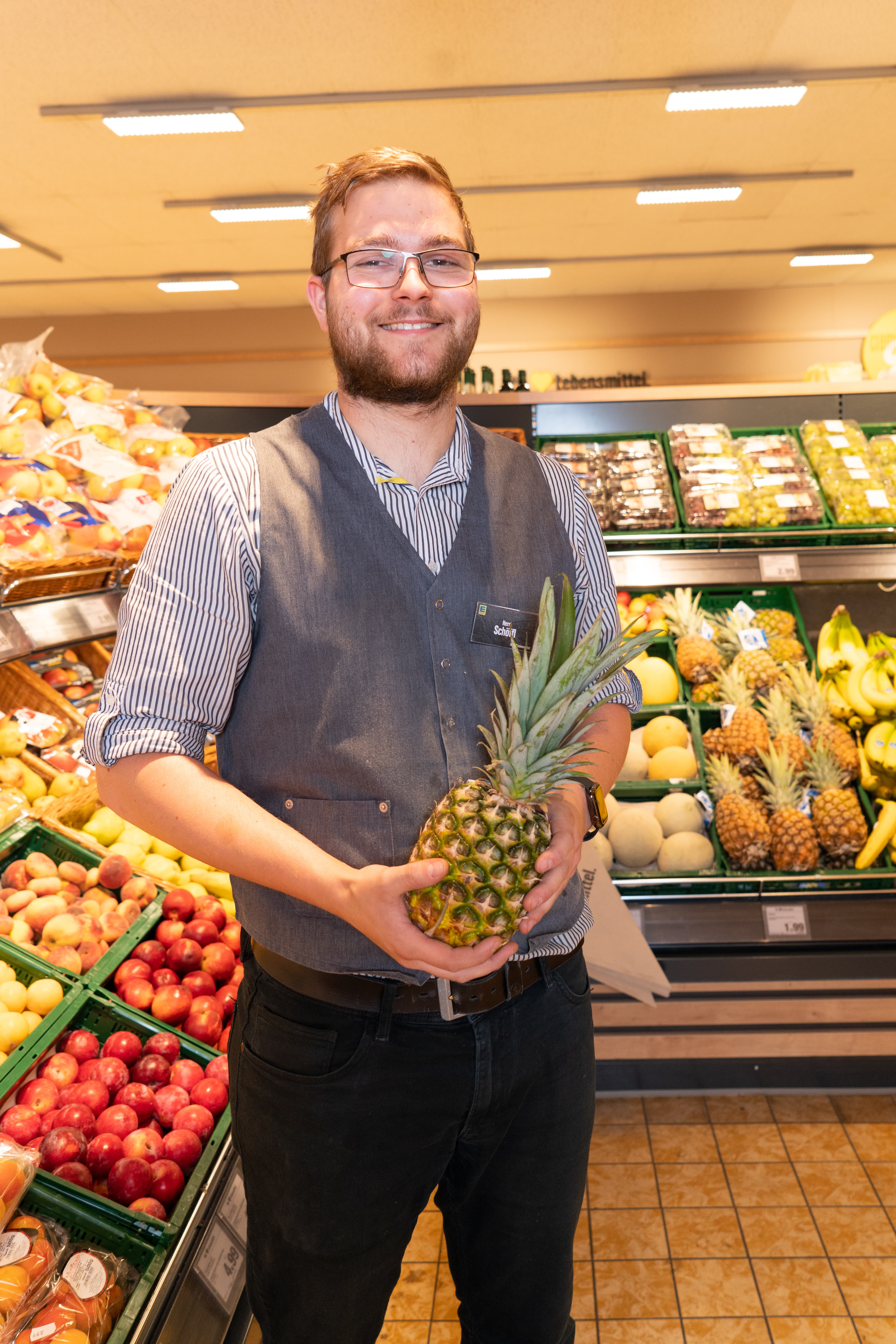 Ein lächelnder Mitarbeiter eines Edeka-Marktes hält eine Ananas in den Händen.