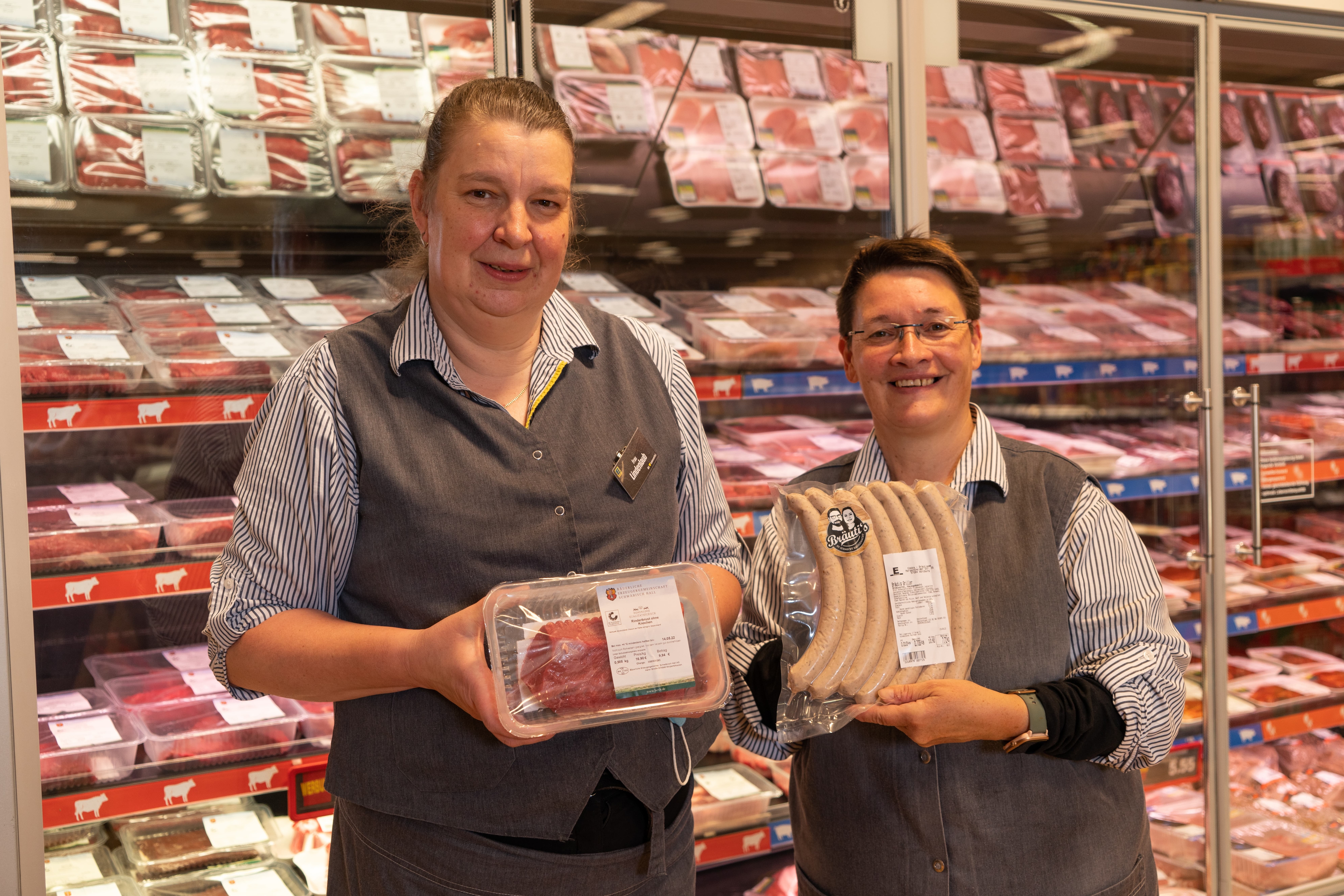 Zwei Mitarbeiterinnen eines Edeka-Marktes. Eine hält ein Stück abgepacktes Fleisch in den Händen. Die andere hält ein Paket Bratwurst in der Hand.