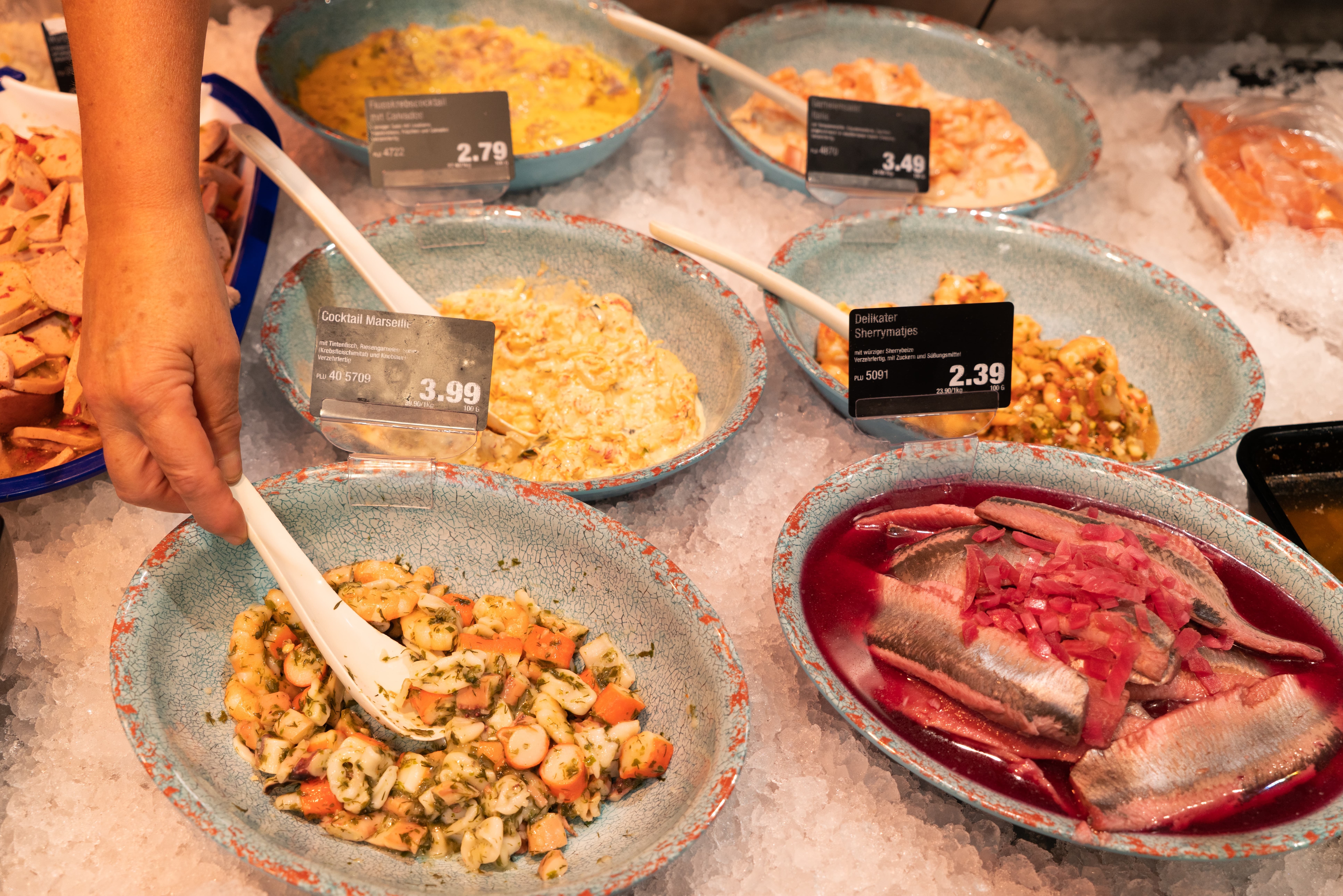 Eine Hand nimmt Meeresfrüchte aus einer Schale. Die Schale steht mit anderen Schalen gefüllt mit Meeresfrüchten in einer Bedientheke.