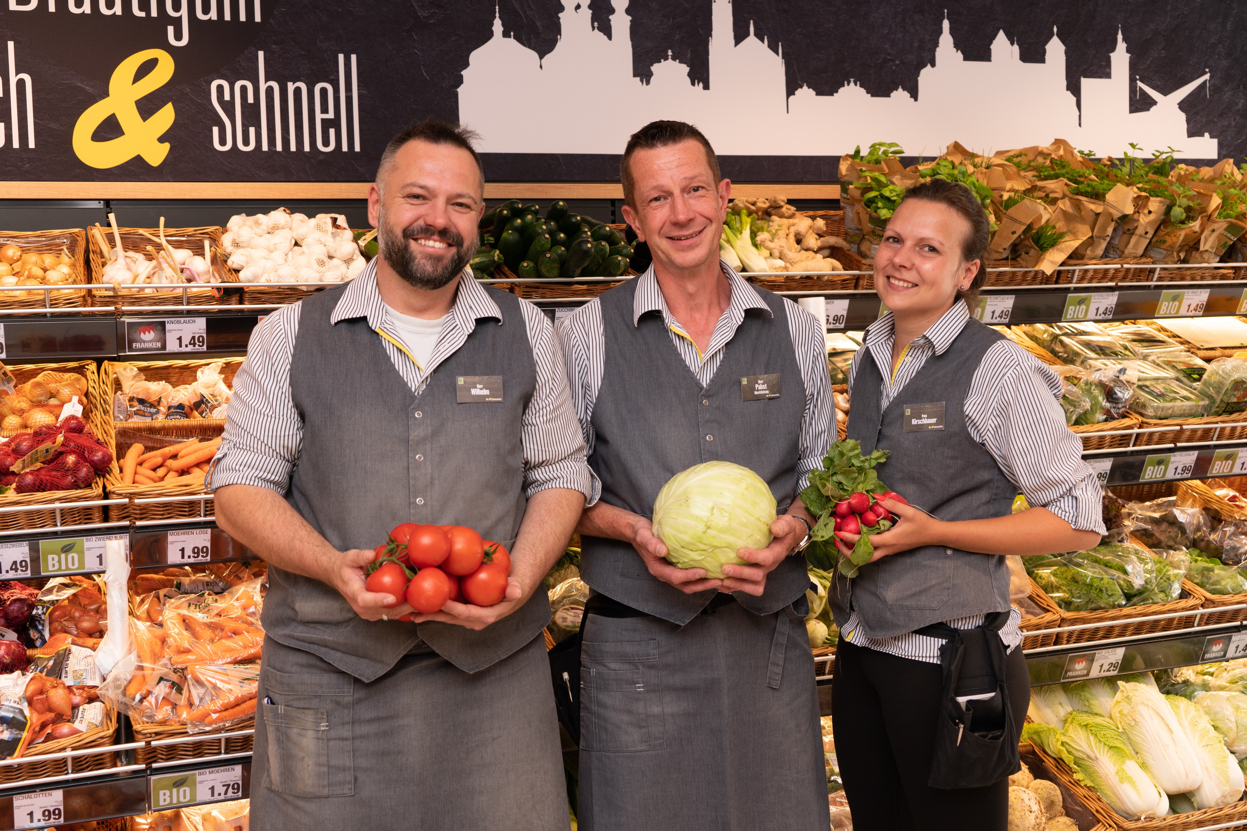 Drei lächelnde Mitarbeitende eines Edeka-Marktes halten verschiedene Sorten Gemüse in den Händen.