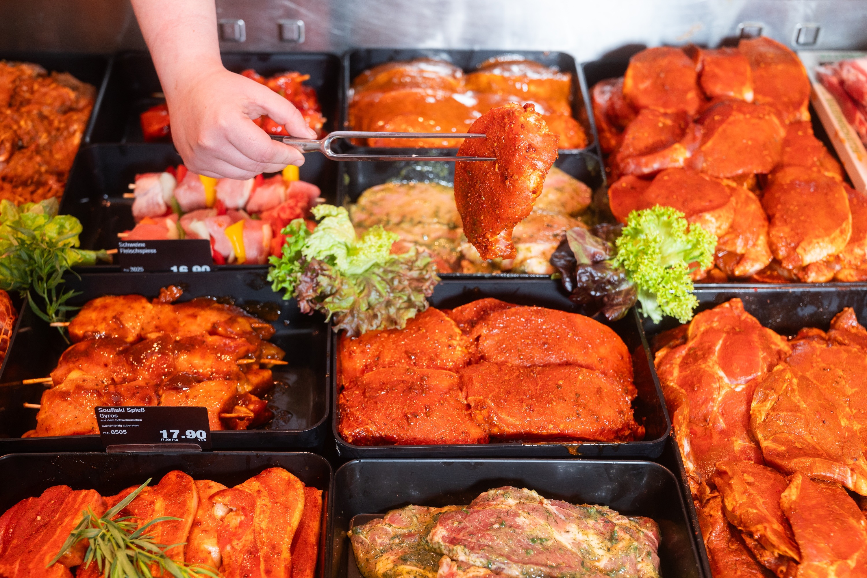 Verschiedene marinierte Fleischstücke. Eine Hand hebt mit einem Spieß ein Stück hoch.