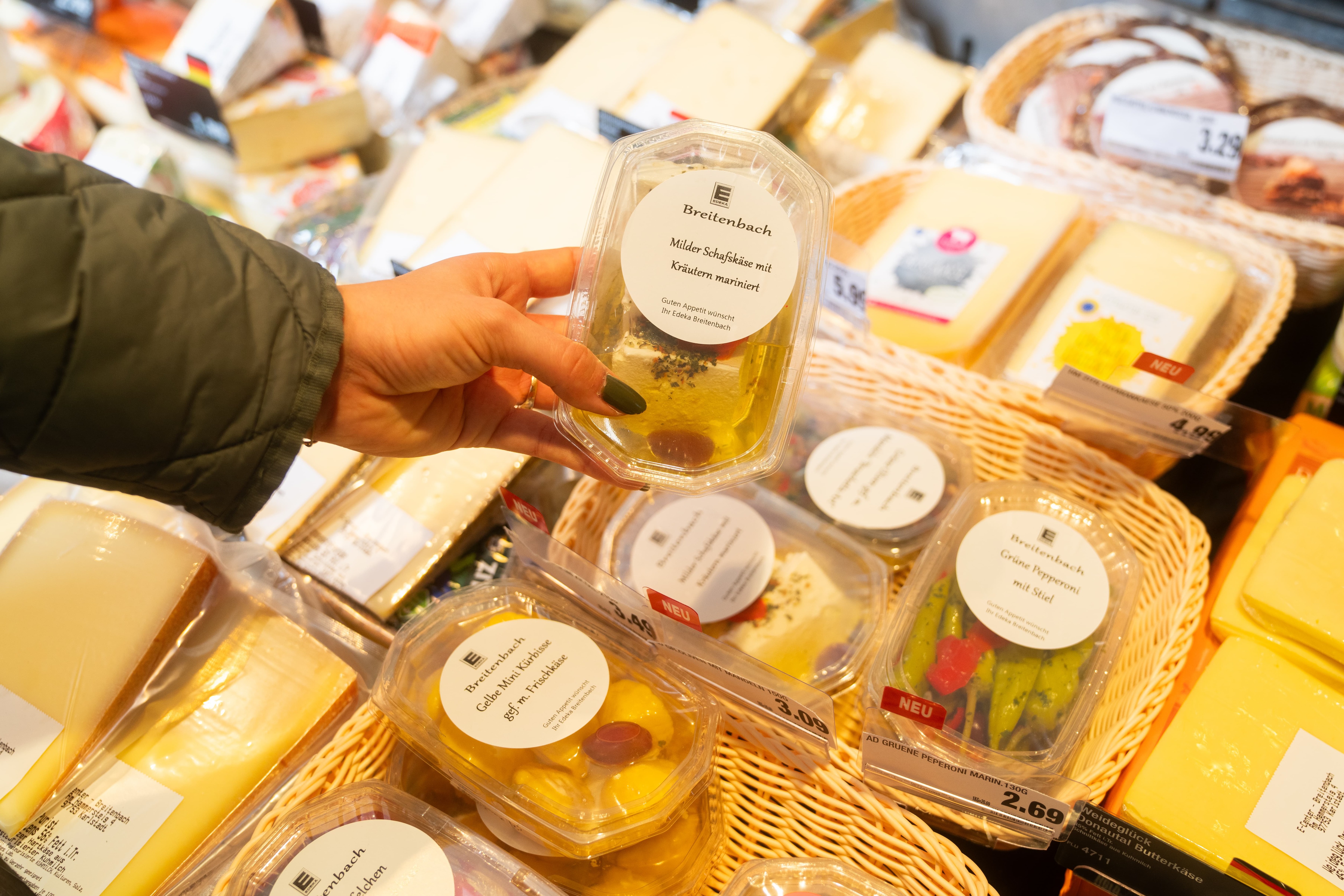 Eine Hand hält eine kleine transparente Plasticpackung hoch, auf der steht: Breitenbach milder Schafskäse mit Kräutern mariniert.