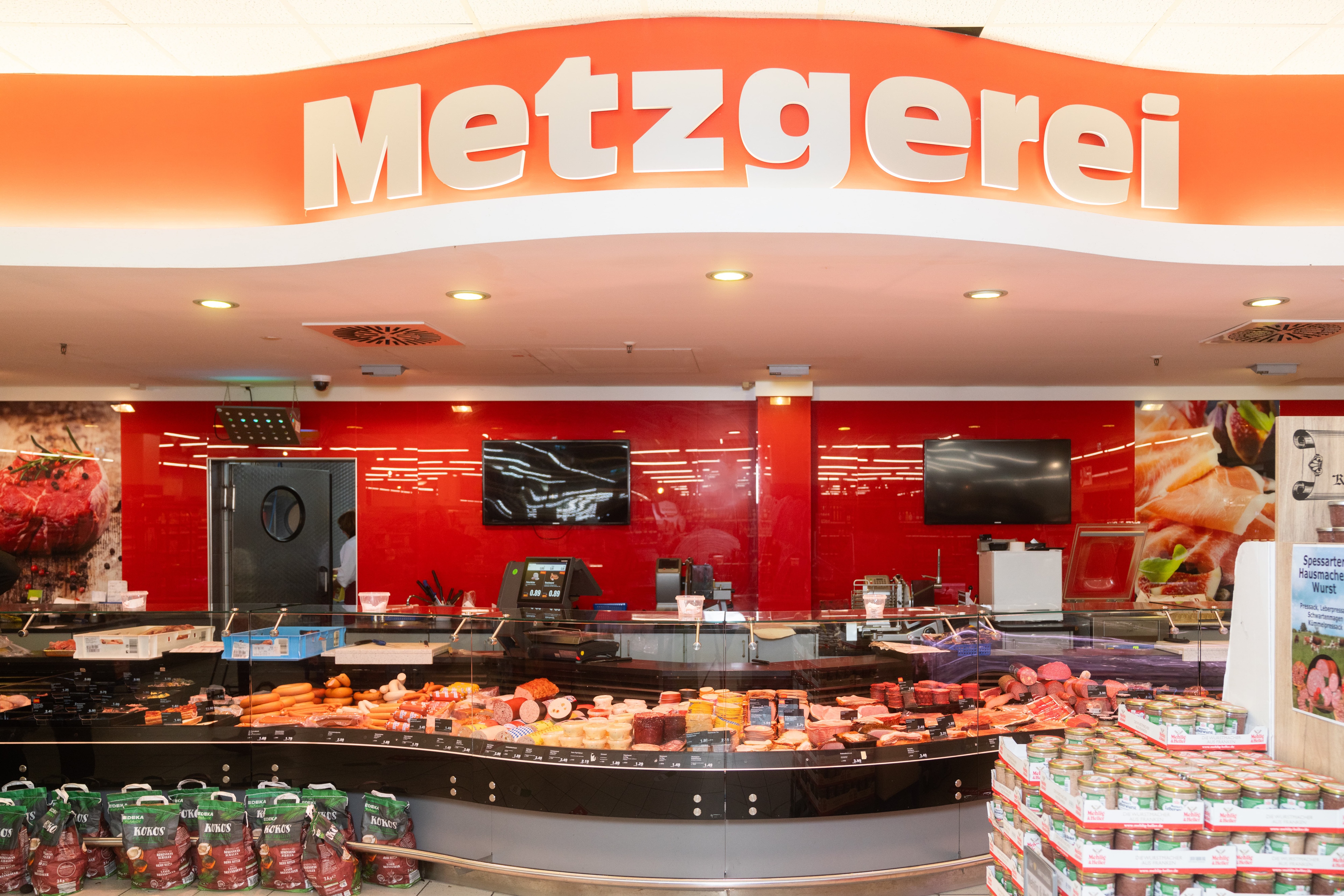 Fleischtheke mit roter Wand und Screens dahinter. Über den Spotlichtern steht Metzgerei.