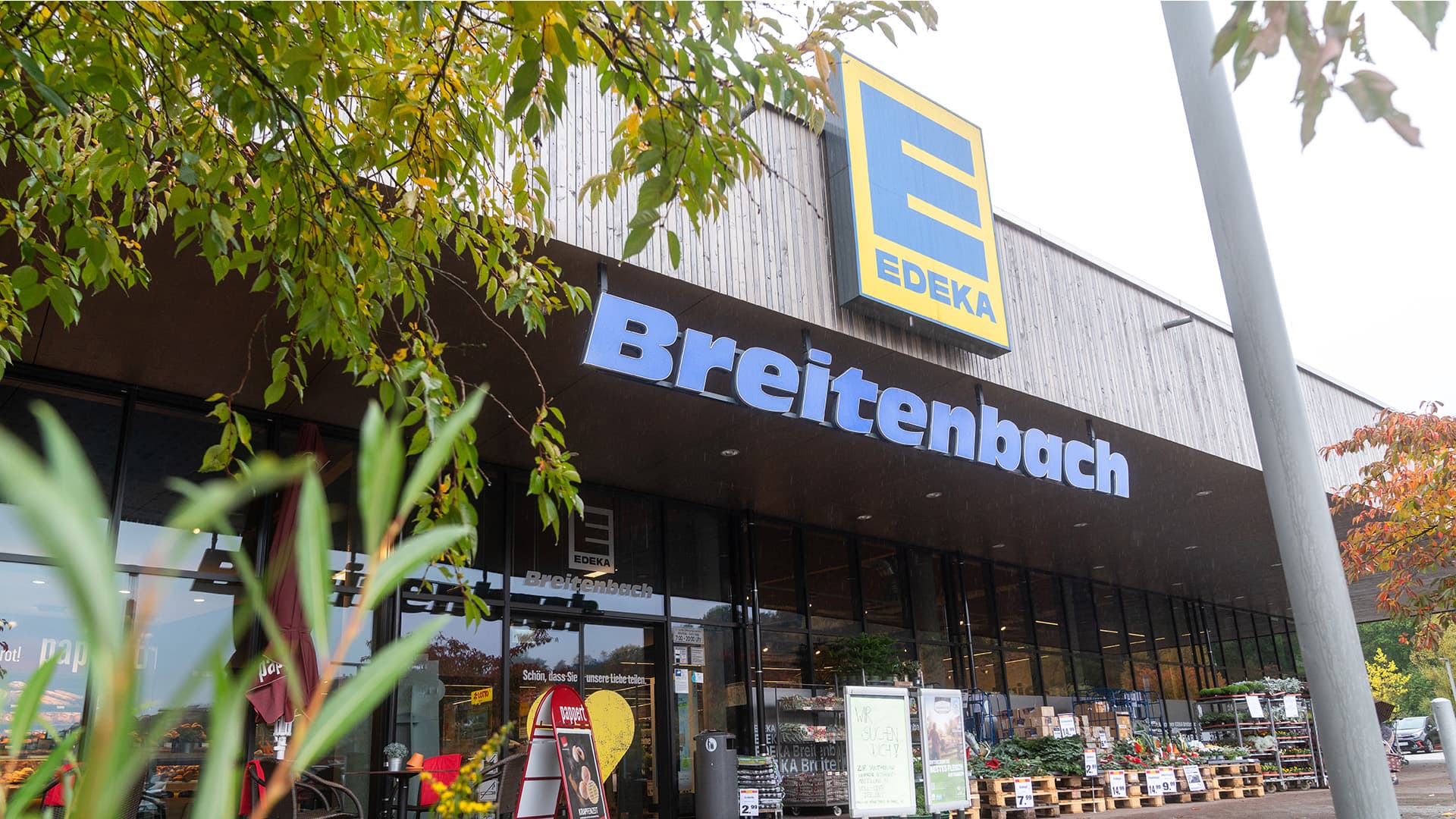 EDEKA Breitenbach vom Parkplatz aus gesehen. Große Glasfassade, davor Paletten mit Pflanzen.