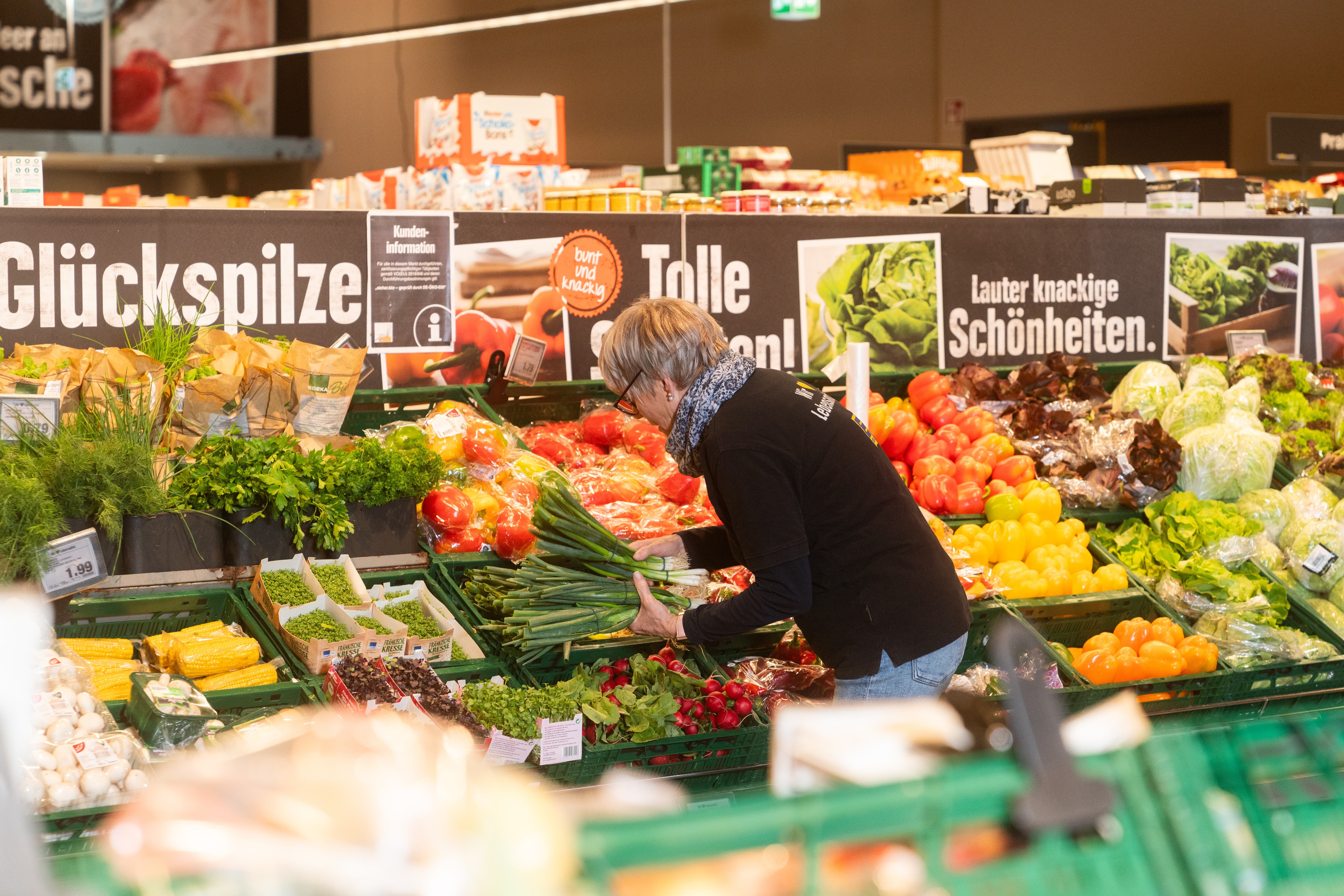 Eine Frau in schwarzem Pullover sortiert Frühlingszwiebeln in einem Gemüseregal ein.
