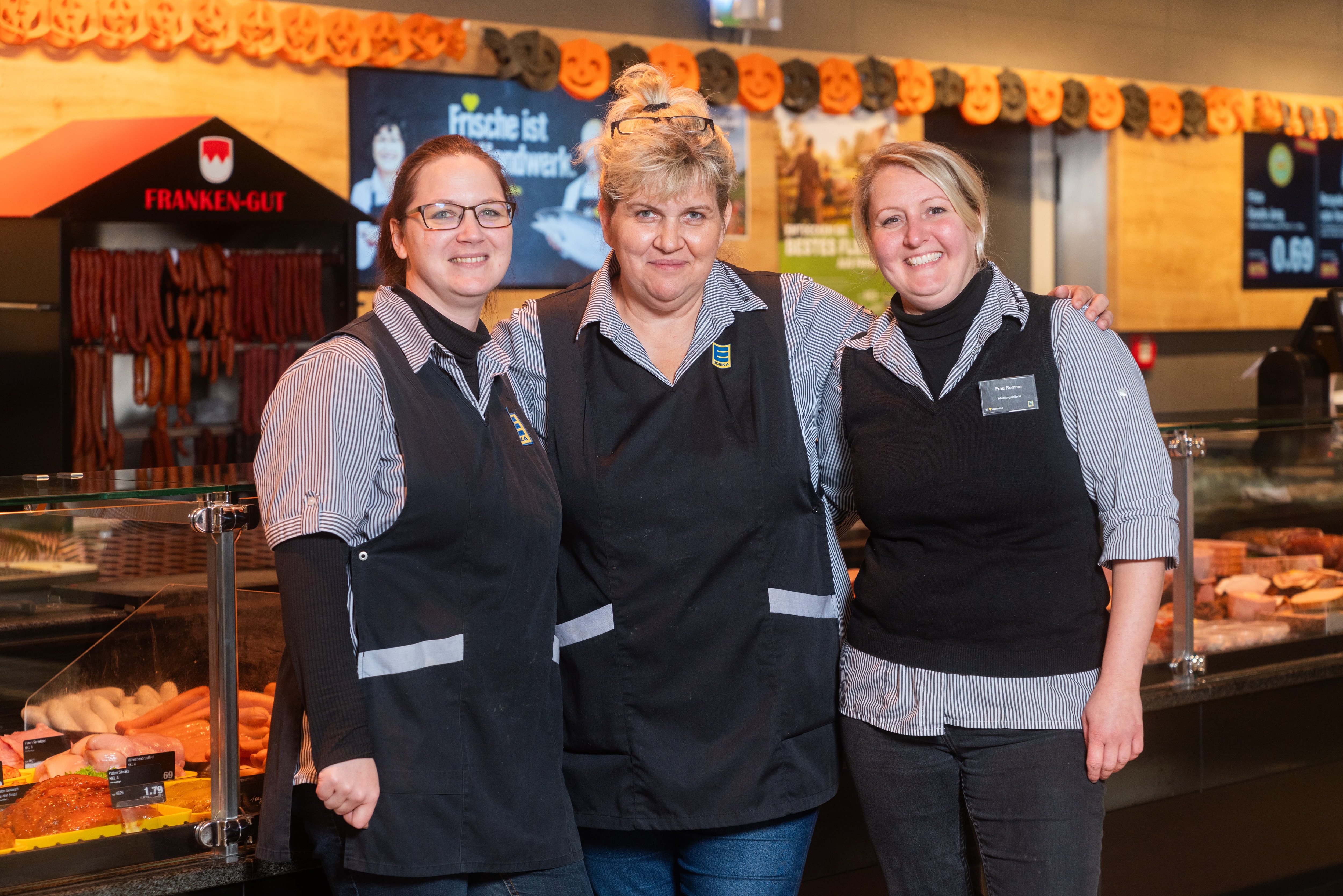 Drei Frauen in Arbeitskleidung von EDEKA stehen lachend vor der Fleischtheke.