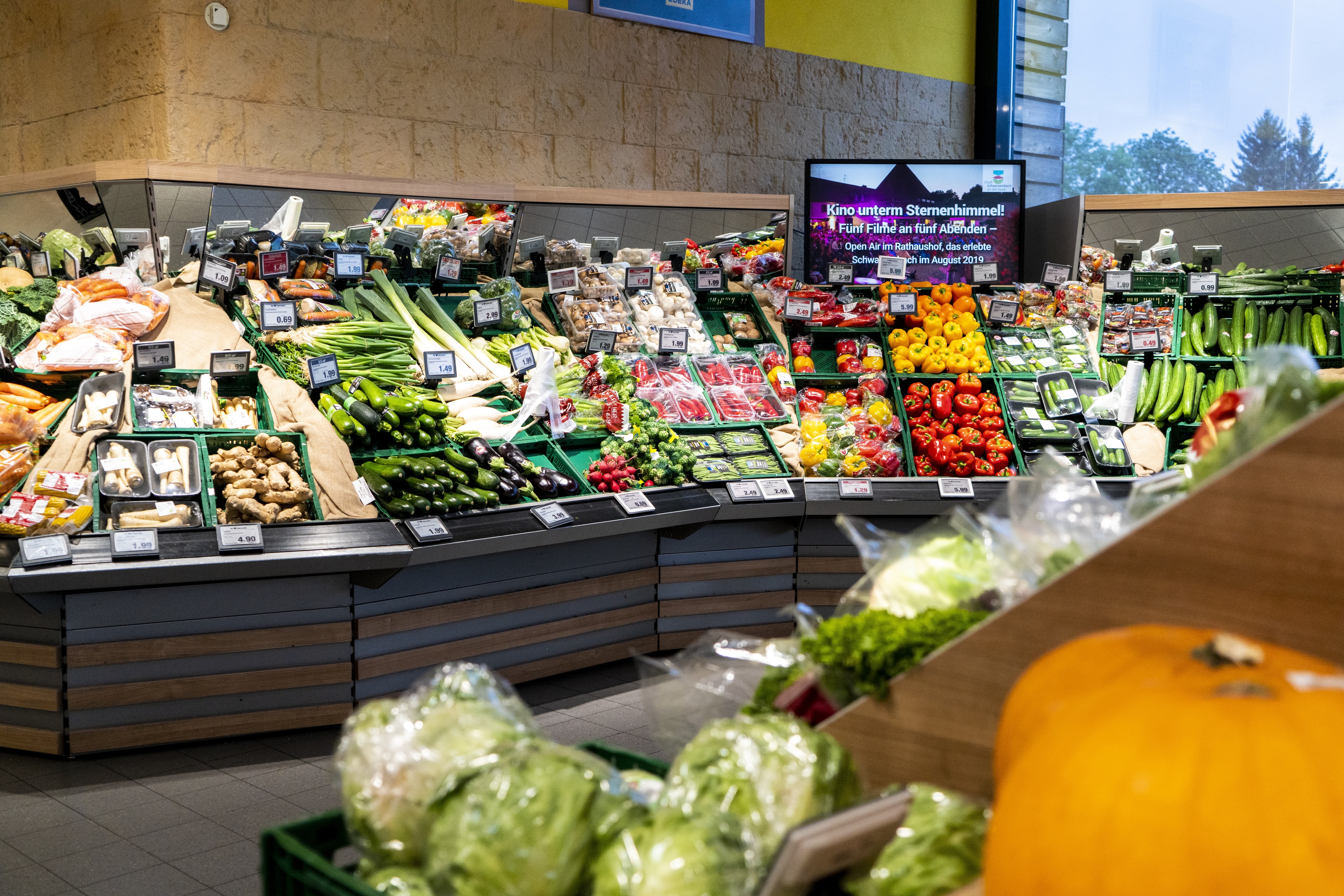Die Gemüseabteilung eines EDEKA-Markts. Über der Paprika ist ein Fernsehbildschirm mit Werbung für ein regionales Event angebracht.