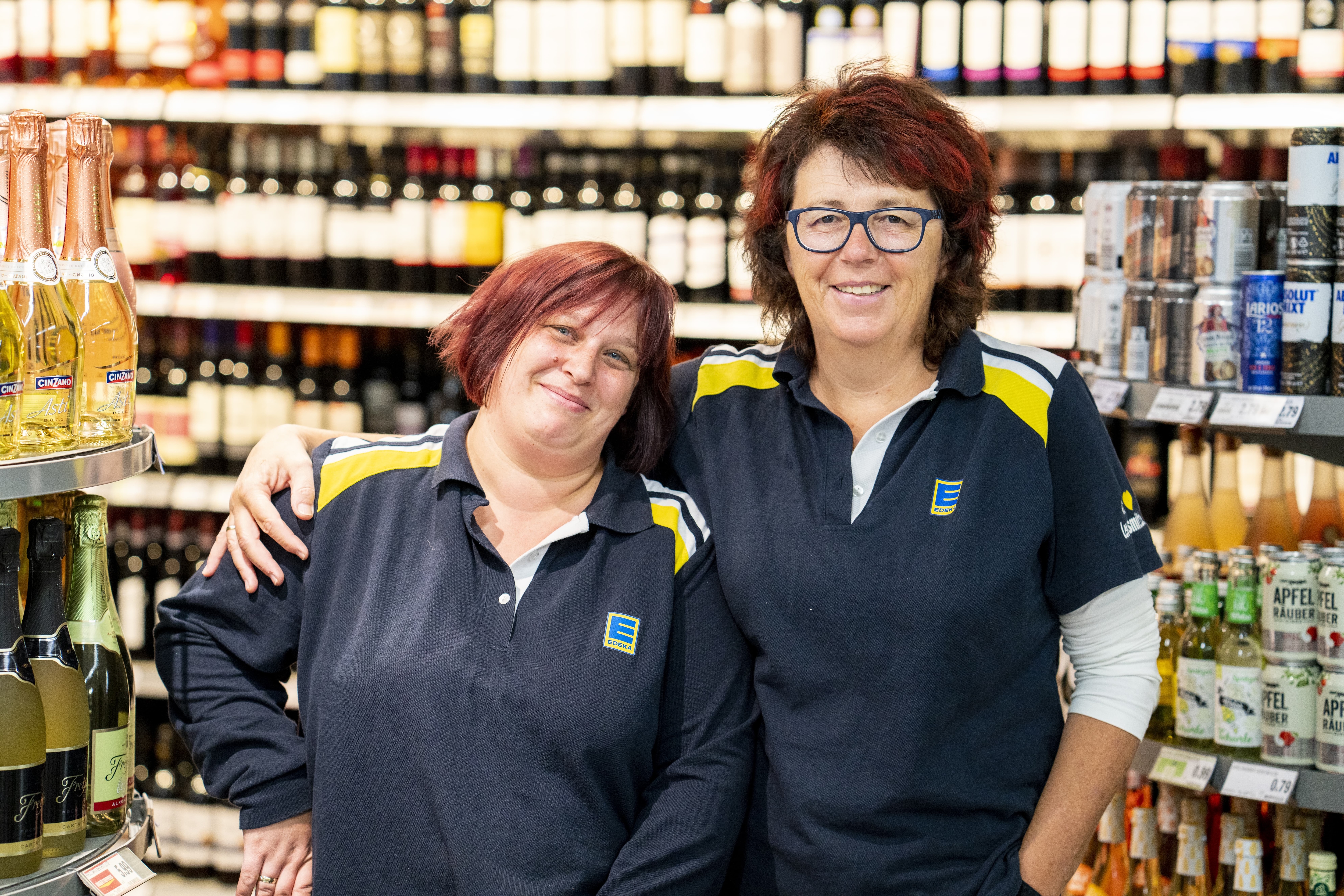 Zwei Mitarbeiterinnen stehen Arm in Arm und lächeln in die Kamera, im Hintergrund Produktregale.