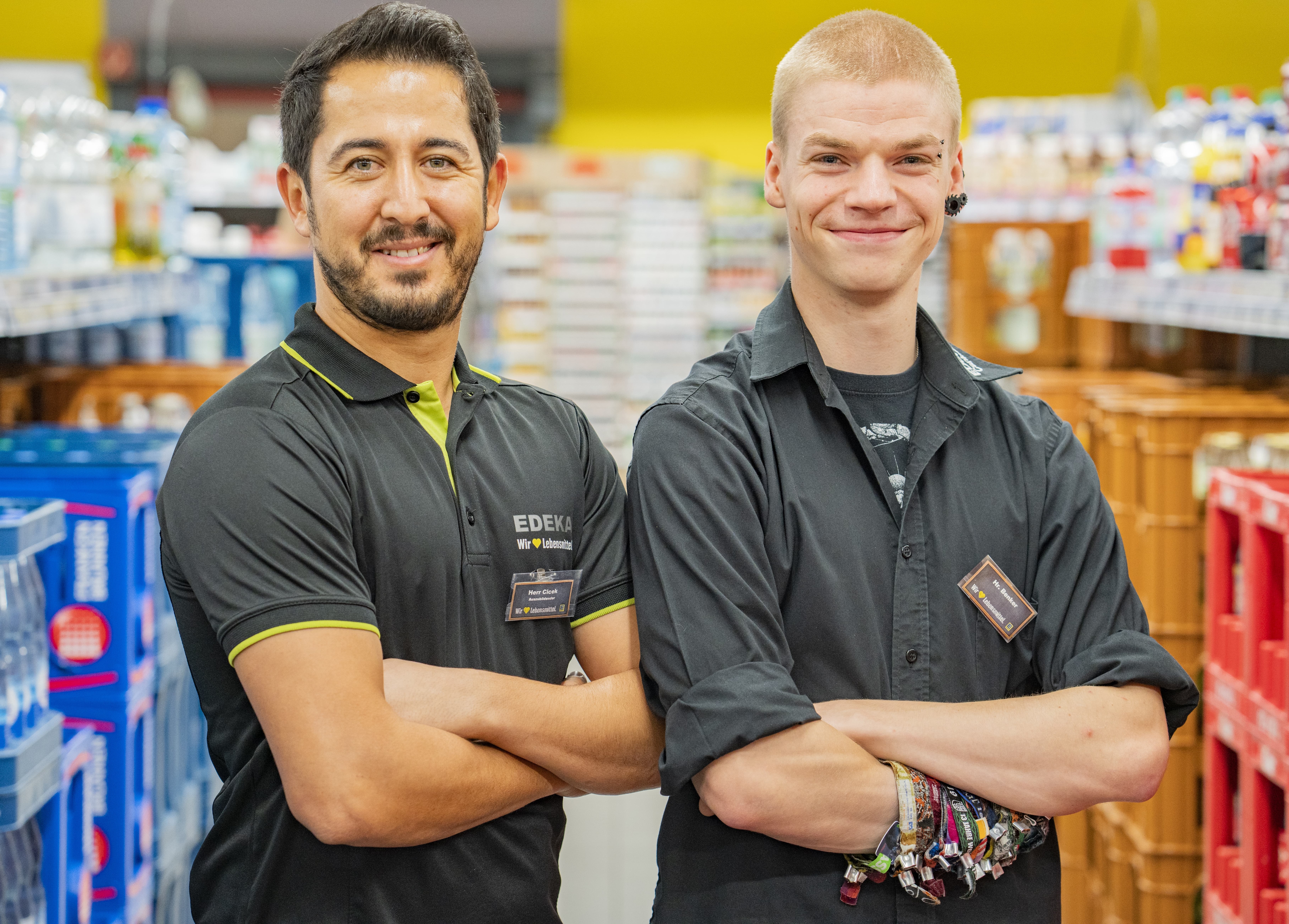 Zwei Mitarbeiter dieses EDEKA-Markts stehen mit verschränkten Armen vor dem Getränkesortiment des Markts und lächeln in die Kamera.