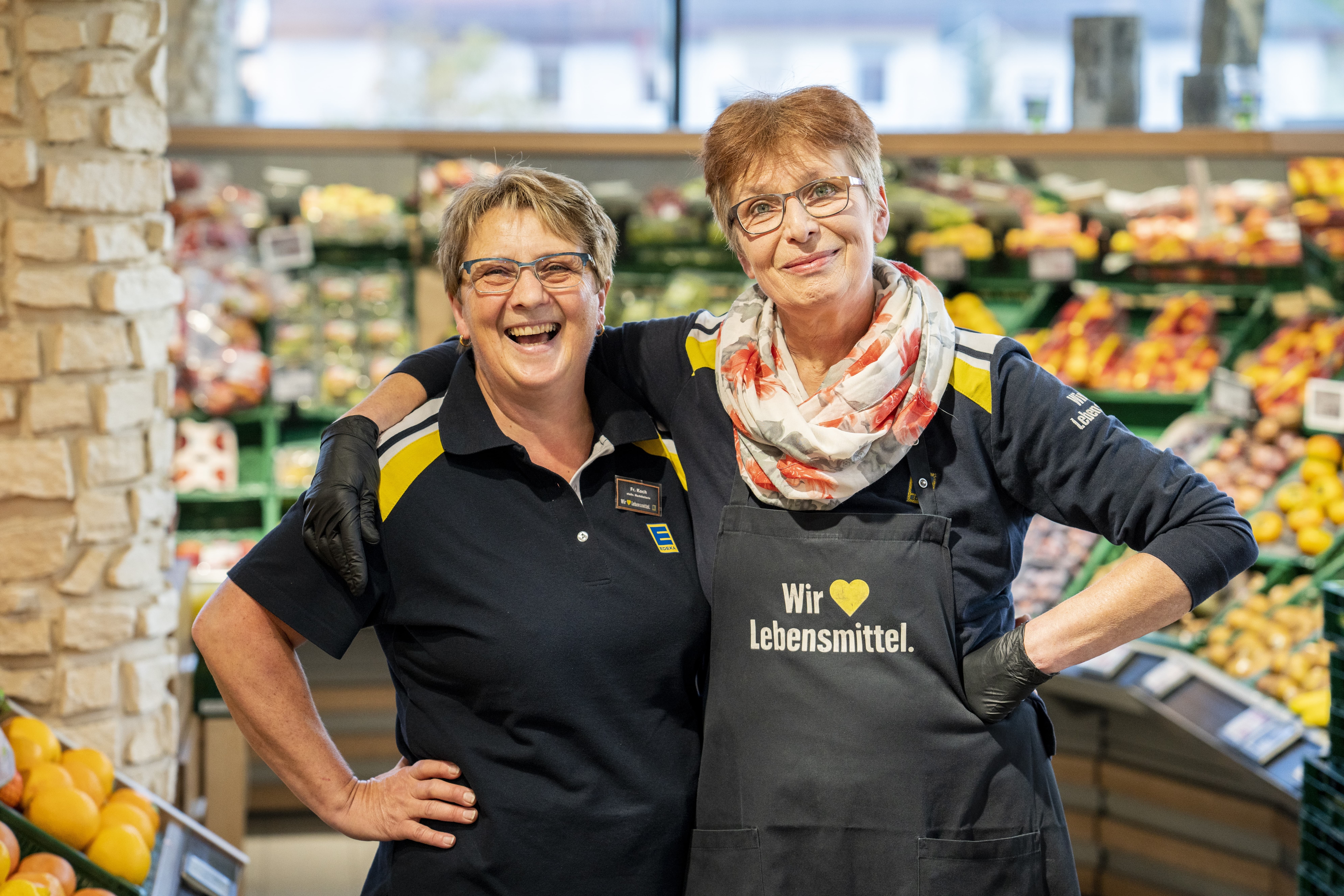 Zwei Mitarbeiterinnen stehen Arm in Arm und lachen in die Kamera, im Hintergrund die Obst- und Gemüseabteilung.