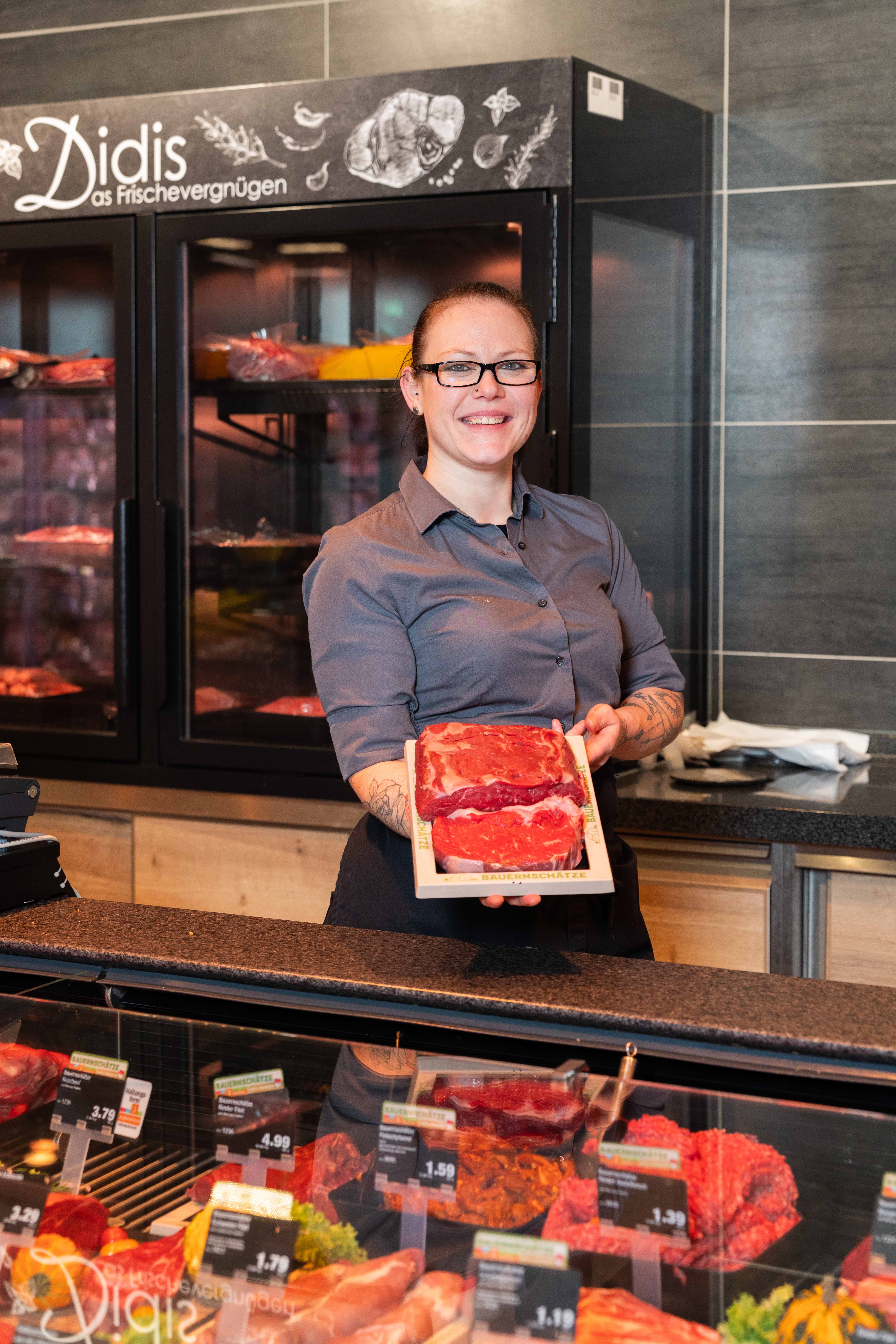 Eine Edeka Mitarbeiterin steht hinter der Fleischtheke. Sie präsentiert zwei Stücke Fleisch, die auf einem Holzbrett liegen.