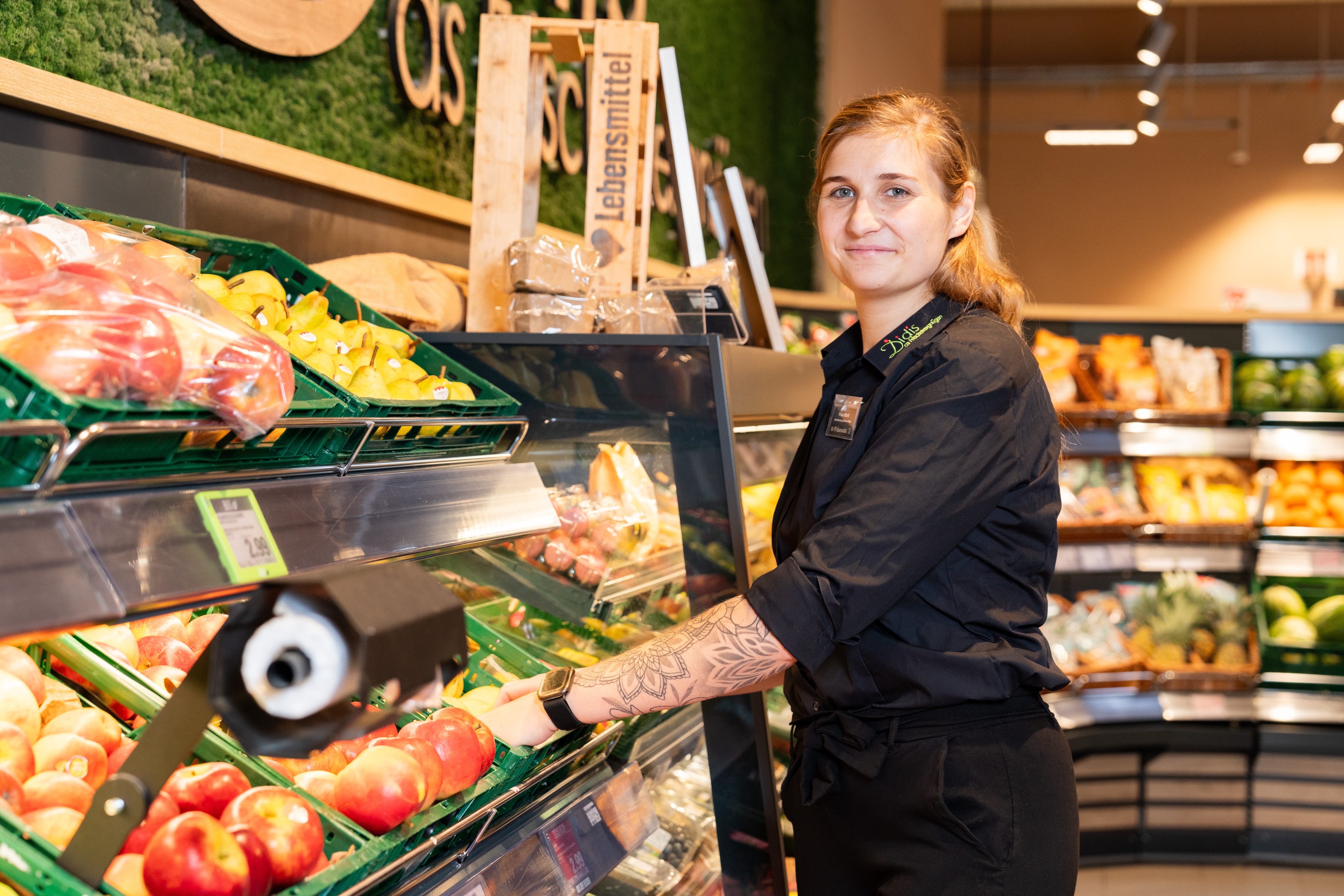 Eine Edeka Mitarbeiterin steht seitlich gewandt in der Obst- und Gemüseabteilung und legt neue Ware in einen Obstkasten.
