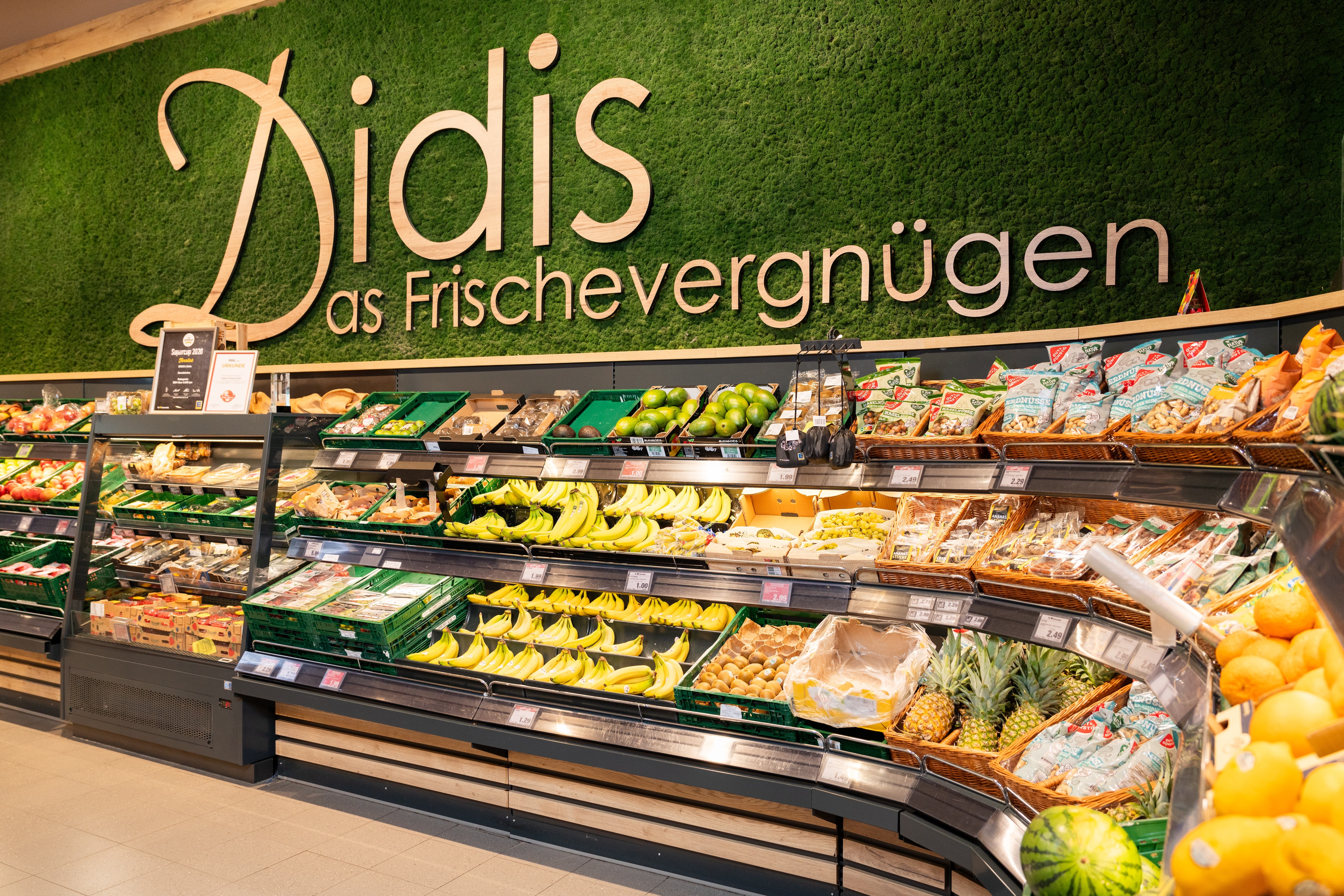 Die Obst- und Gemüseabteilung eines EDEKA-Markts mit unterschiedlichen Obst zum Beispiel Bananen und Äpfeln. Über dem Obst ist ein Schriftzug mit der Aufschrift "Didis - Das Frischevergnügen" zu sehen