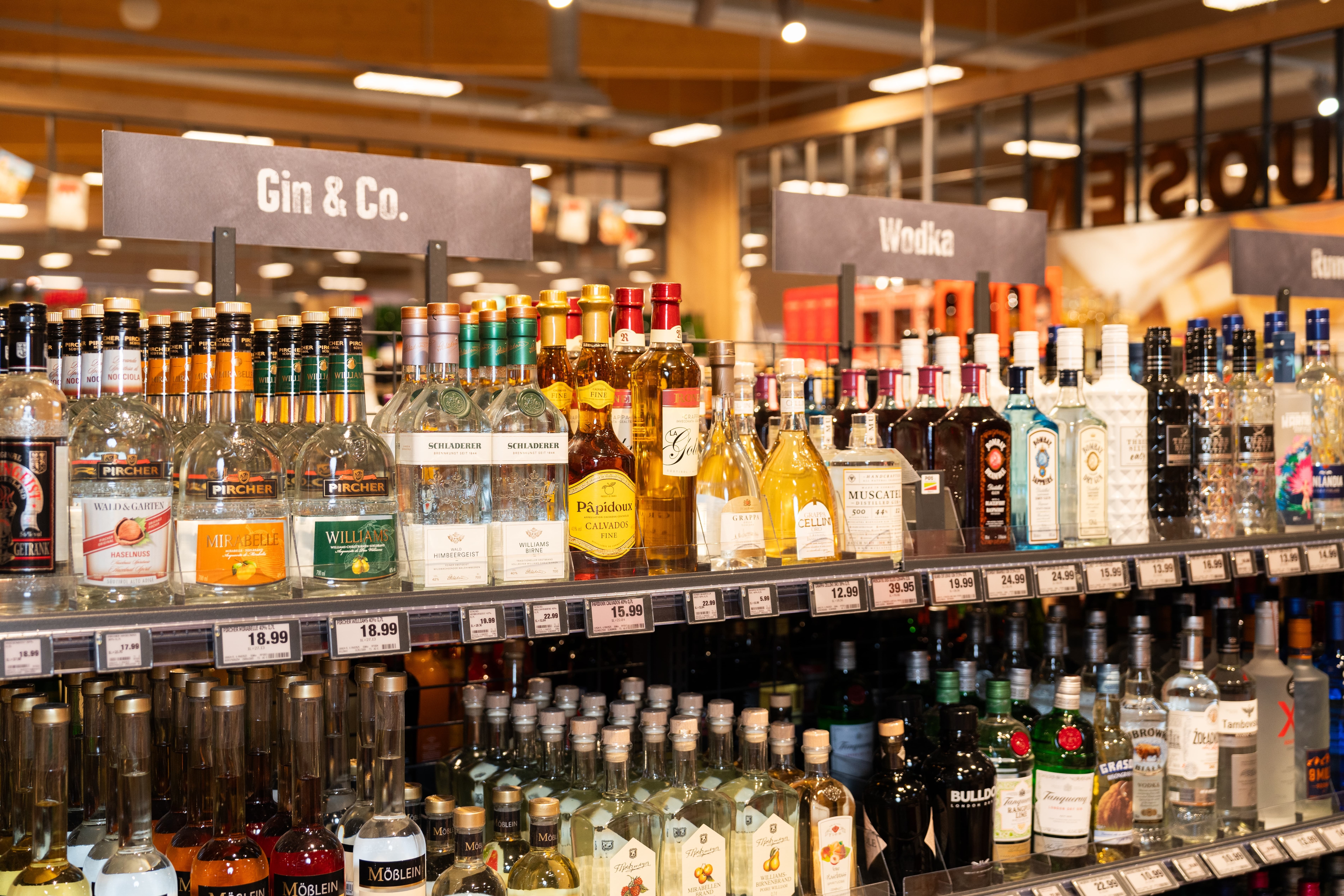 Unterschiedliche Spirituosen aufgereiht in einem Regal. Es sind Schilder mit der Aufschrift "Gin & Co" und "Wodka" im Hintergund zu erkennen.