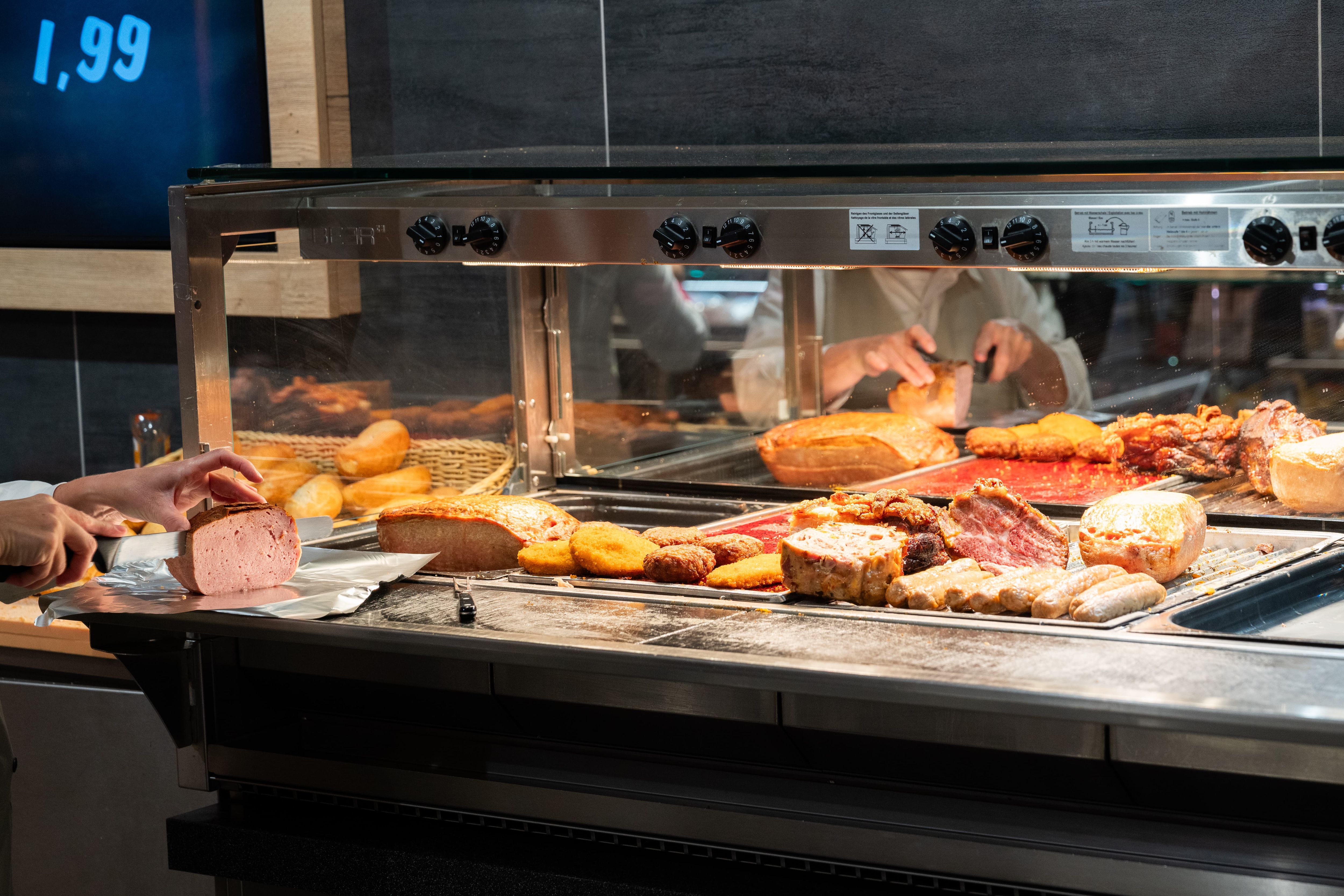 Verschiedene warme Speisen in der Warmen Theke. Eine Mitarbeiterin schneidet ein Stück eines Leberkäses ab.