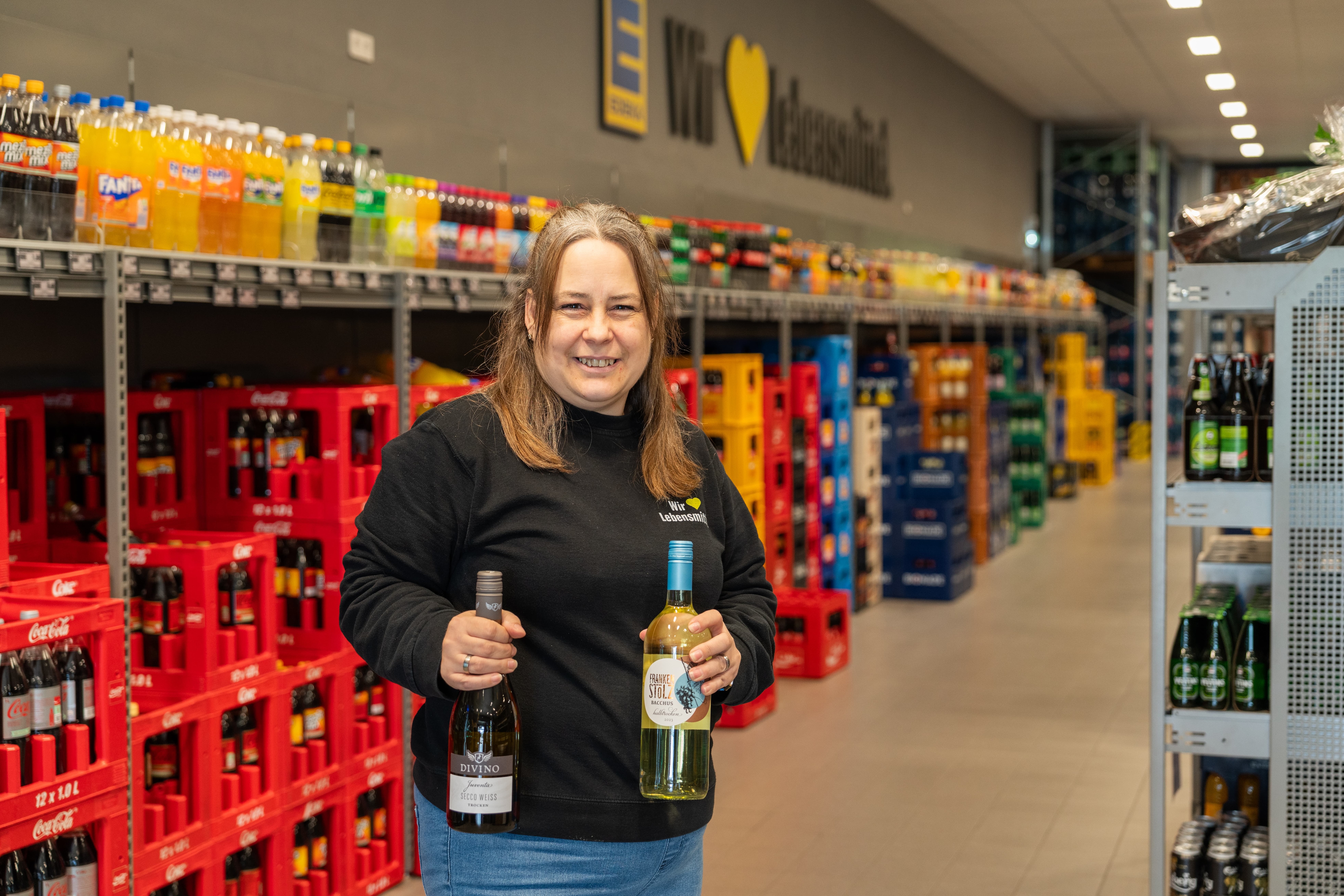 Eine lächelnde Mitarbeiterin steht in einem Getränkemarkt und hält zwei Flaschen Wein in den Händen.
