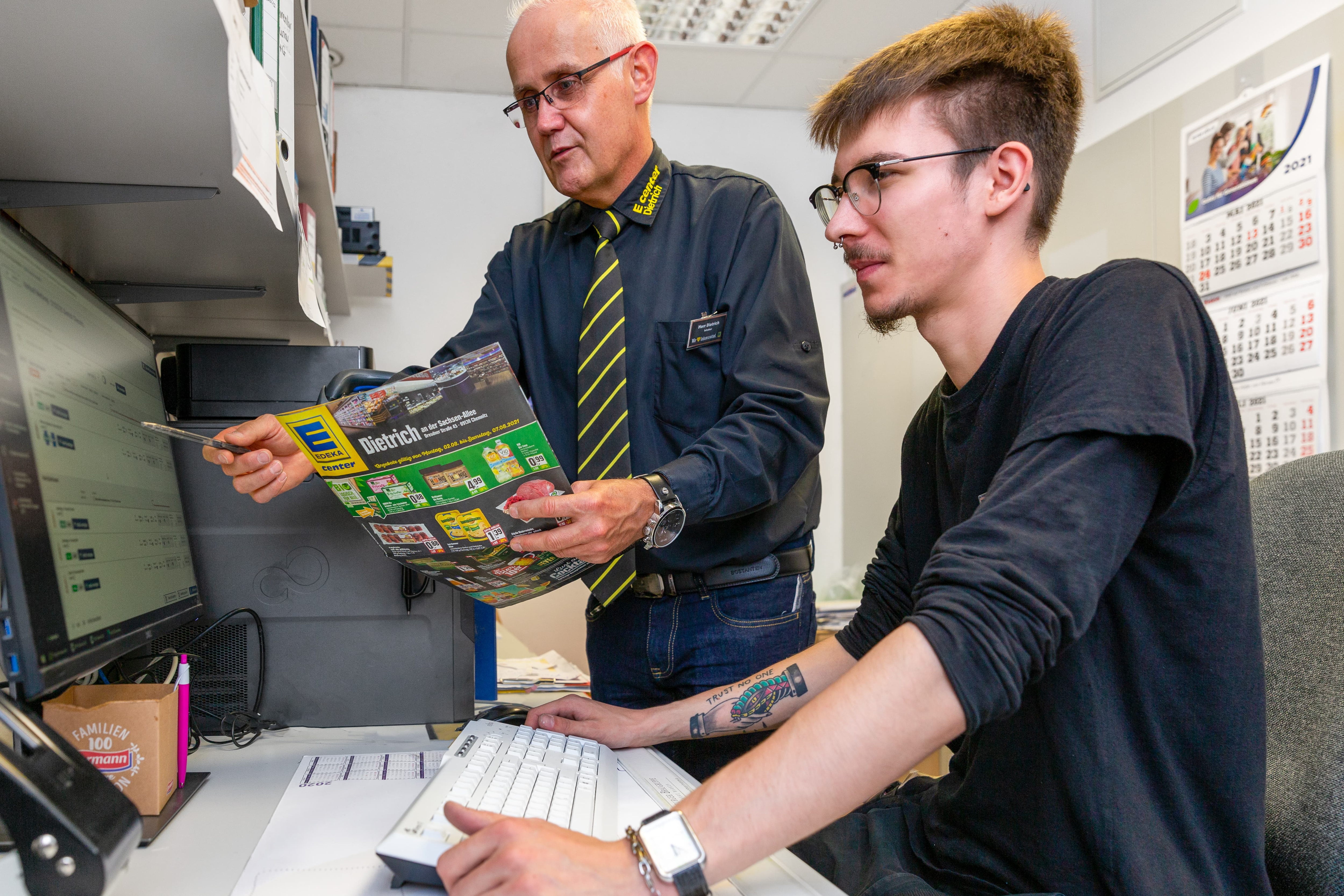 Der Marktleiter erklärt einem Auszubildenden etwas am PC in einem Büro.
