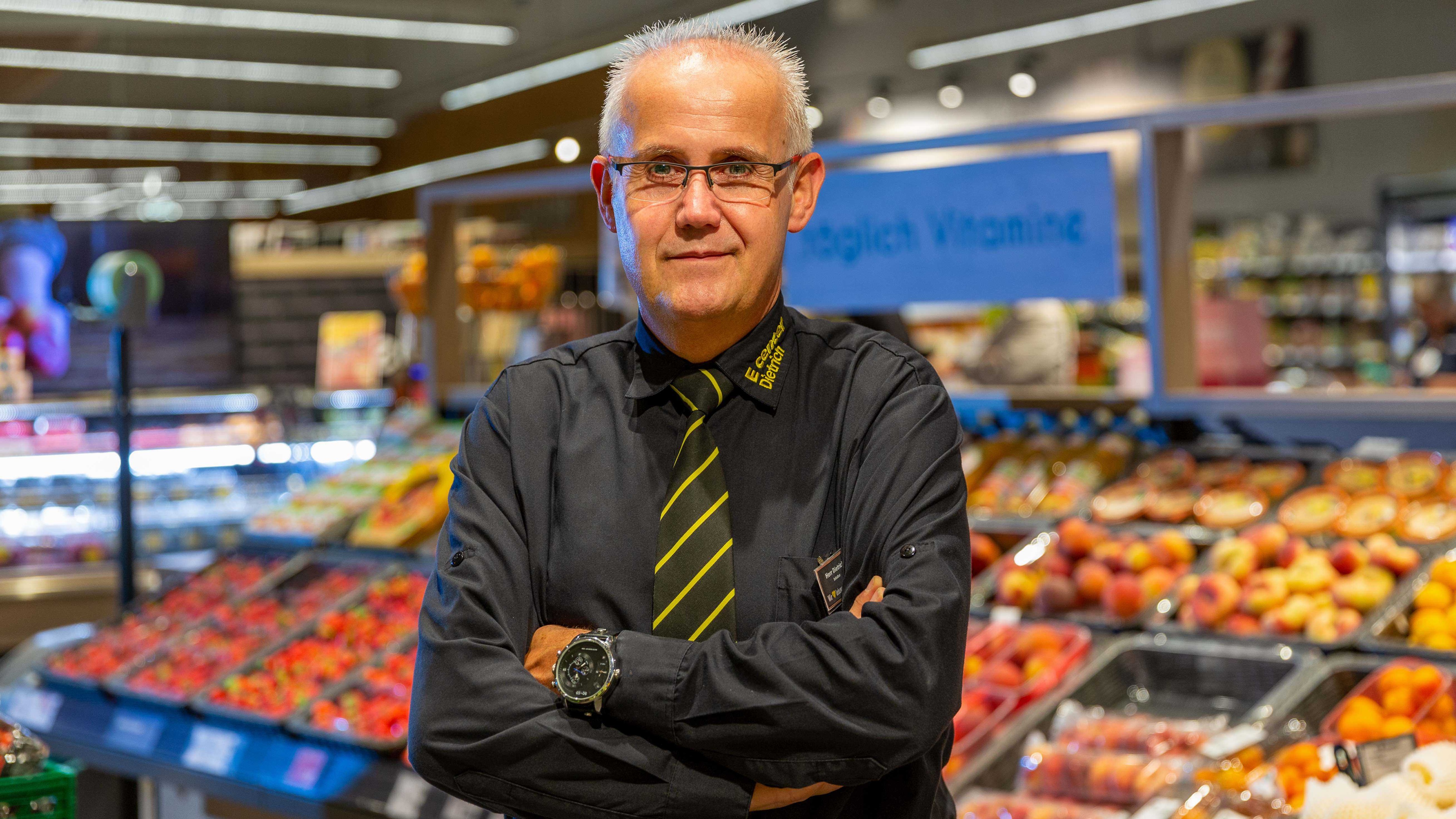 Der Marktinhaber steht in der Obst- und Gemüseabteilung mit einem kleinen Lächeln und überkreuzten Armen.