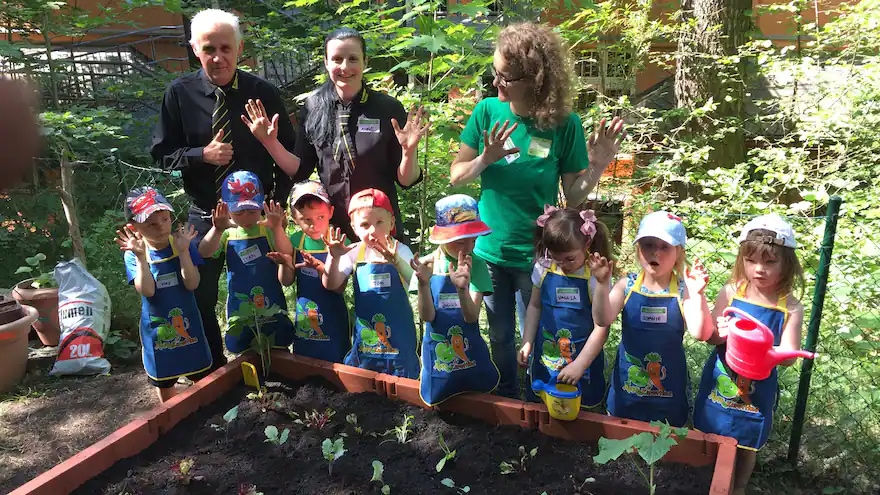 Einige Kinder mit Schürze und Mütze halten die Hände hoch und stehen vor einem Hochbeet gemeinsam mit dem Marktinhaber und zwei Frauen.