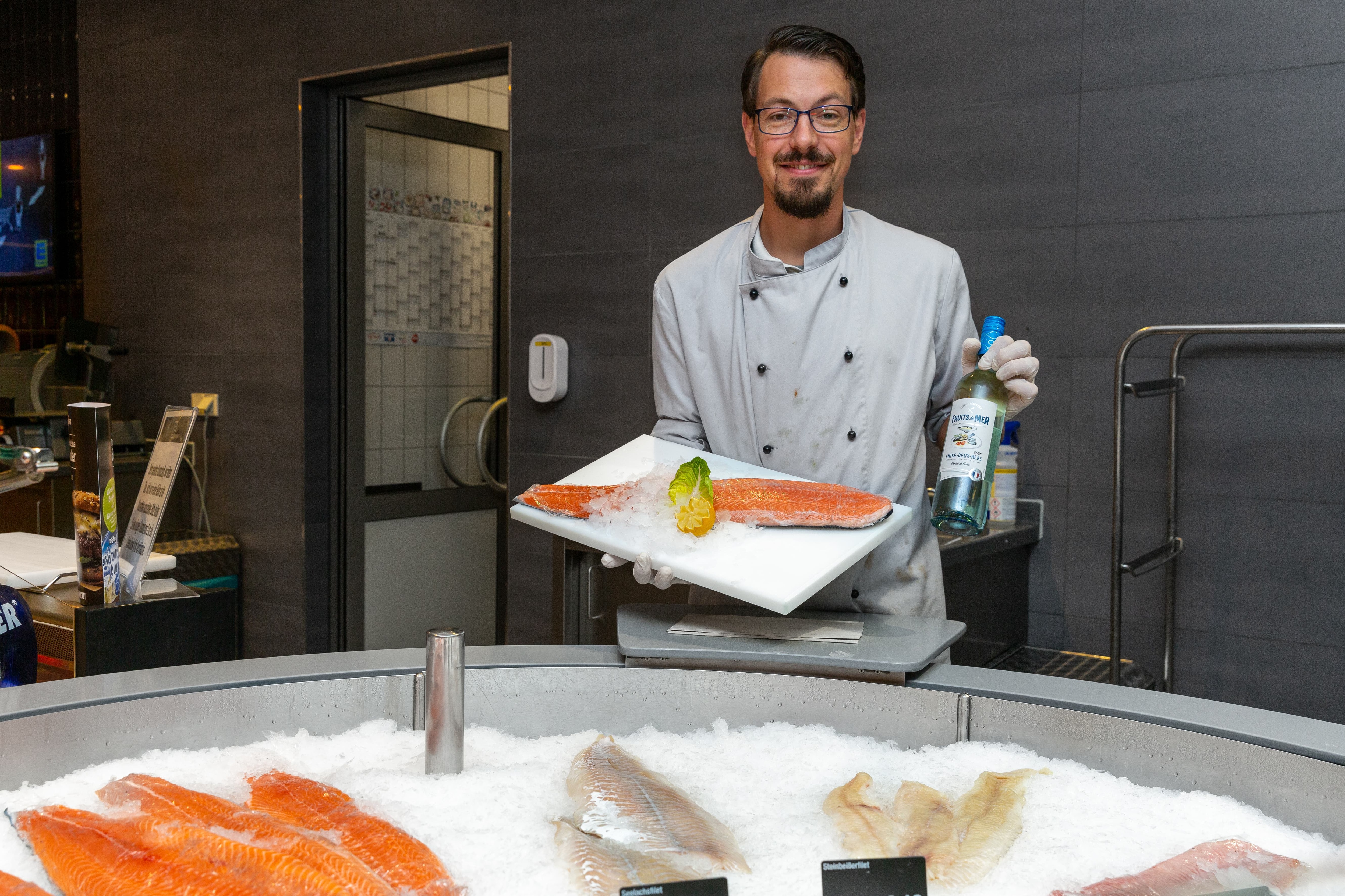 Ein Mitarbeiter hält in der einen Hand ein Brett mit einem Stück Fisch drauf und in der anderen Hand eine Flasche Wein.