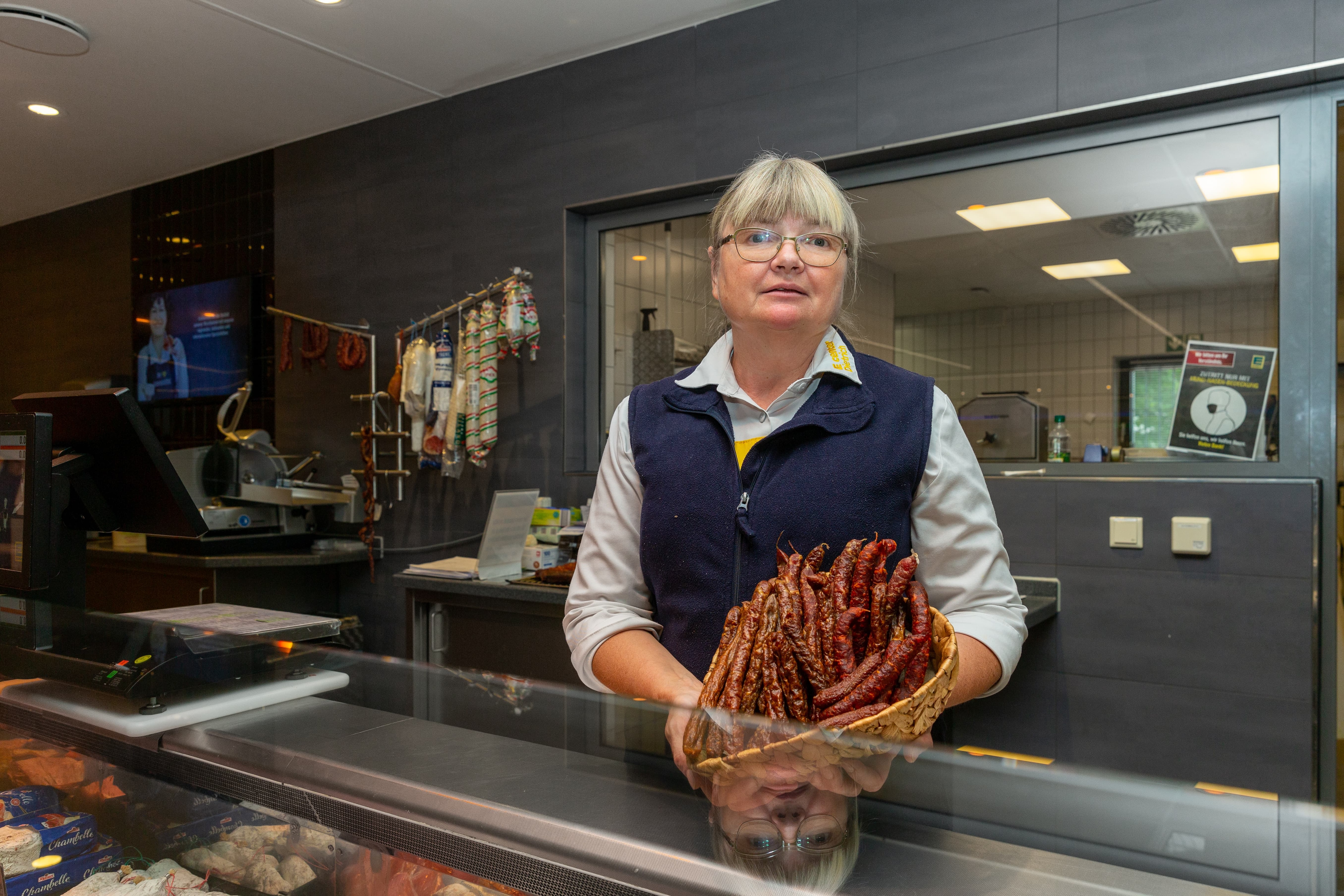 Eine Mitarbeiterin steht hinter der Servicetheke und hält einen Korb mit verschiedenen Würsten.
