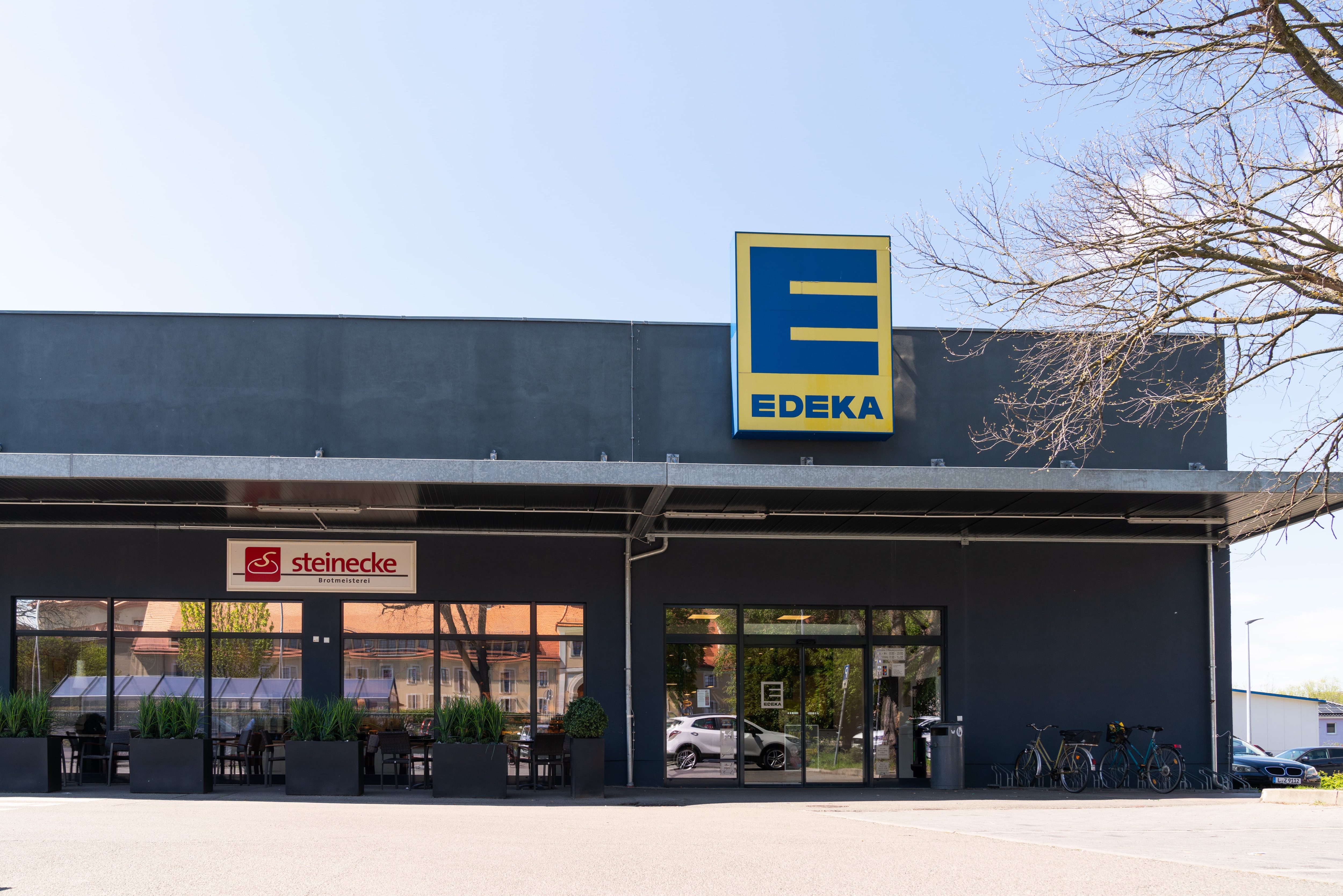 Fassade mit Glastür eines EDEKA-Marktes. In der Eingangstür spiegelt sich ein Auto.