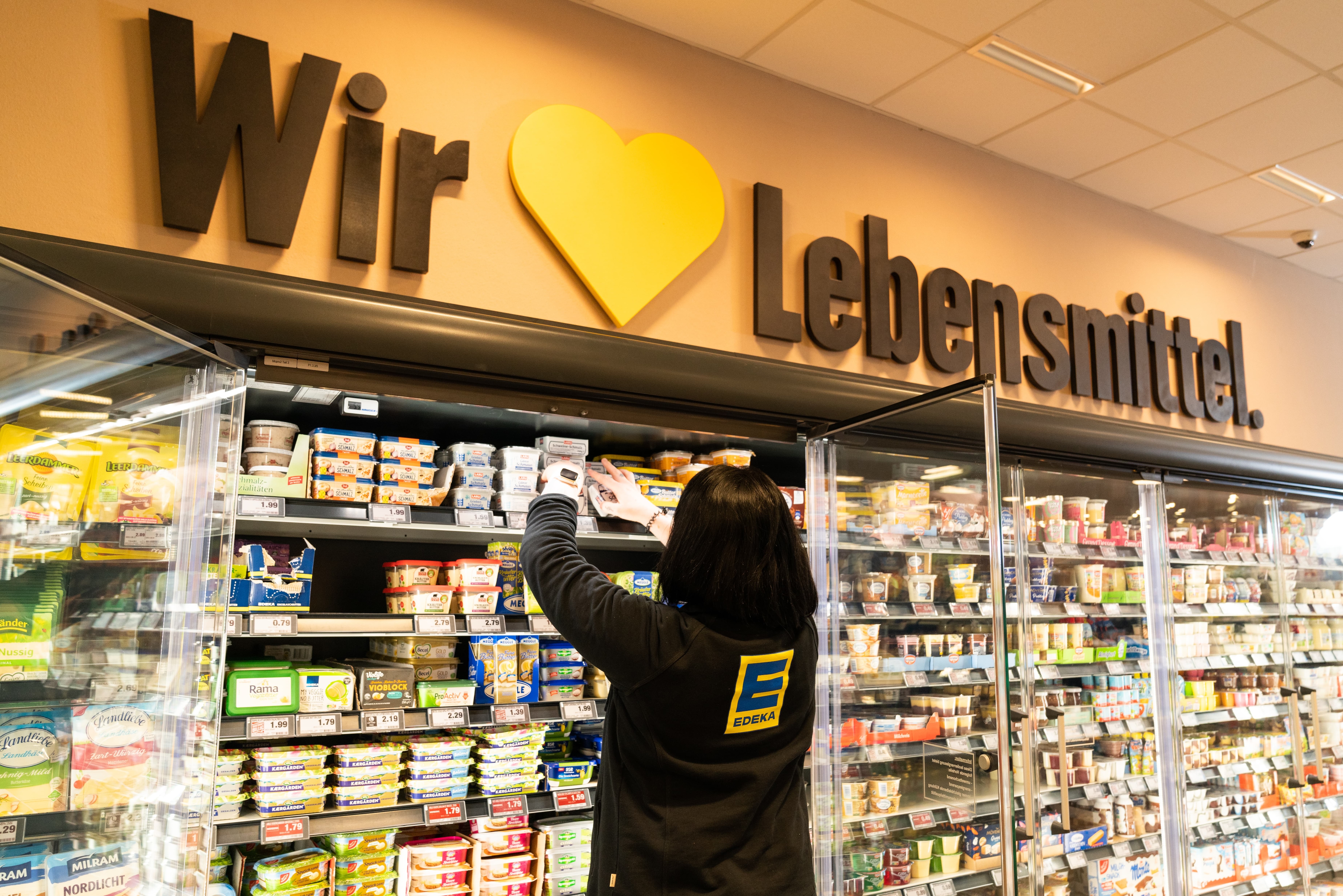 Ein Frau in schwarzer EDEKA-Jacke greift in den offenen Kühlschrank für Molkereiprodukte.