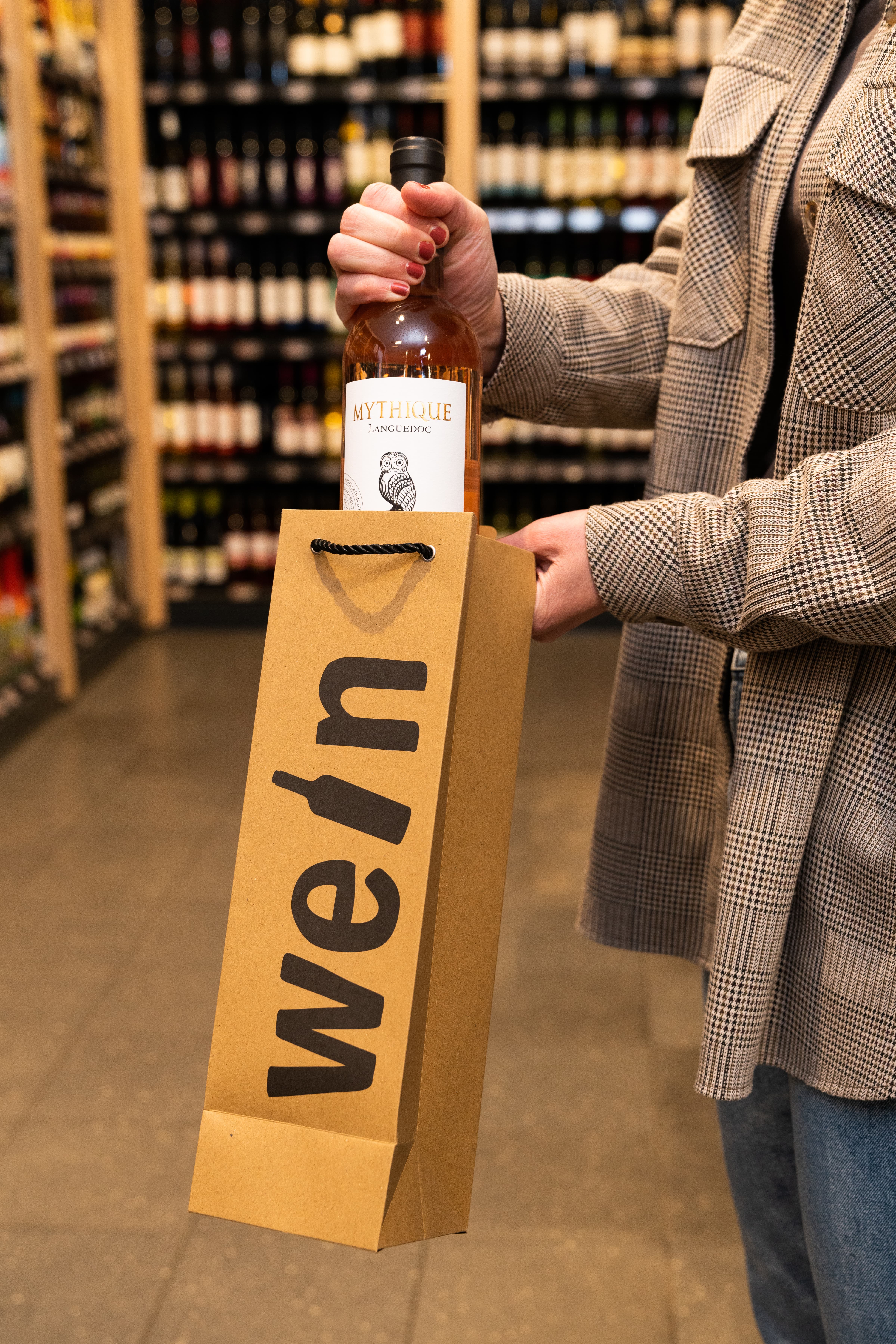 Eine weibliche Person steckt eines Flasche Mythique Languedoc in eine braune Papiertüte mit der Aufschrift Wein.