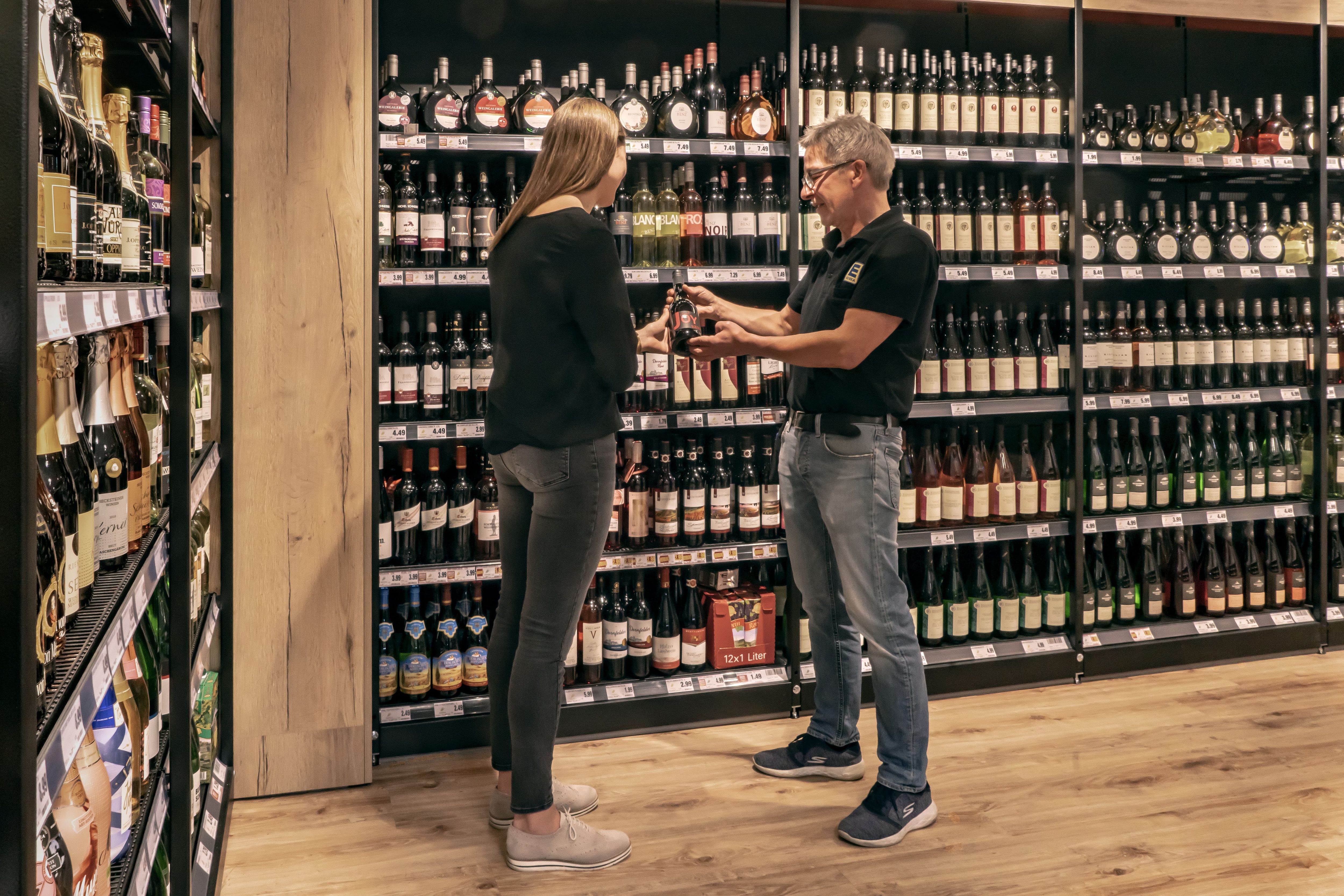 Die Weinabteilung eines EDEKA-Markts. Ein Mitarbeiter zeigt einer Kundin ein Getränk.