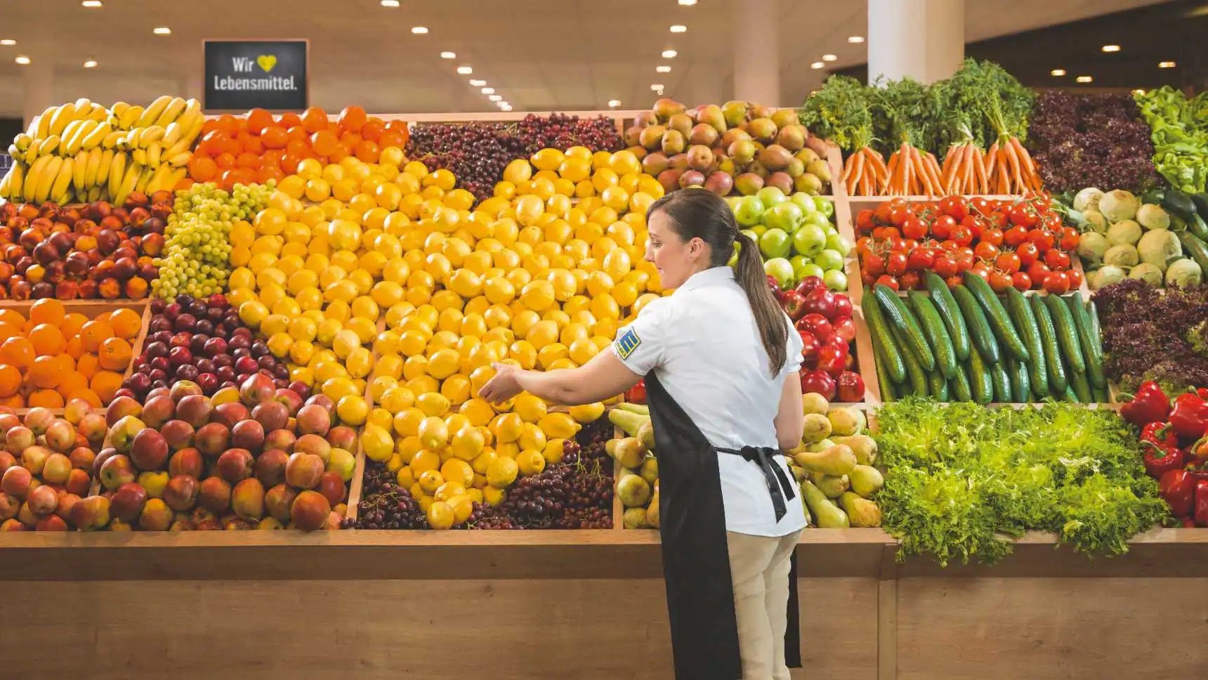 Eine EDEKA-Mitarbeiterin legt ein Herz aus gelben Zitronen in die Auslage einer Obstabteilung.